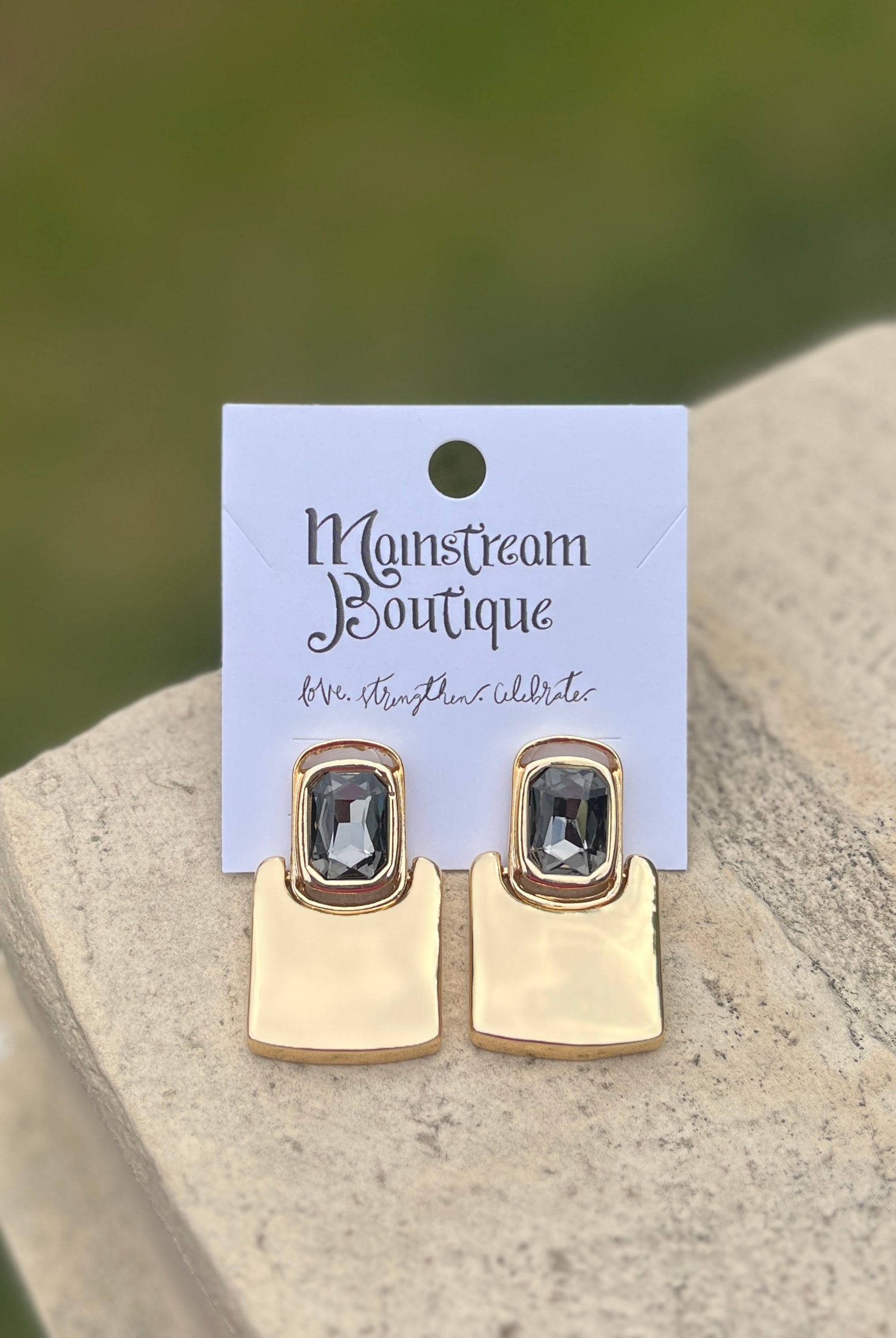 Gold earrings with gemstones placed on a stone surface with a blurred green background.