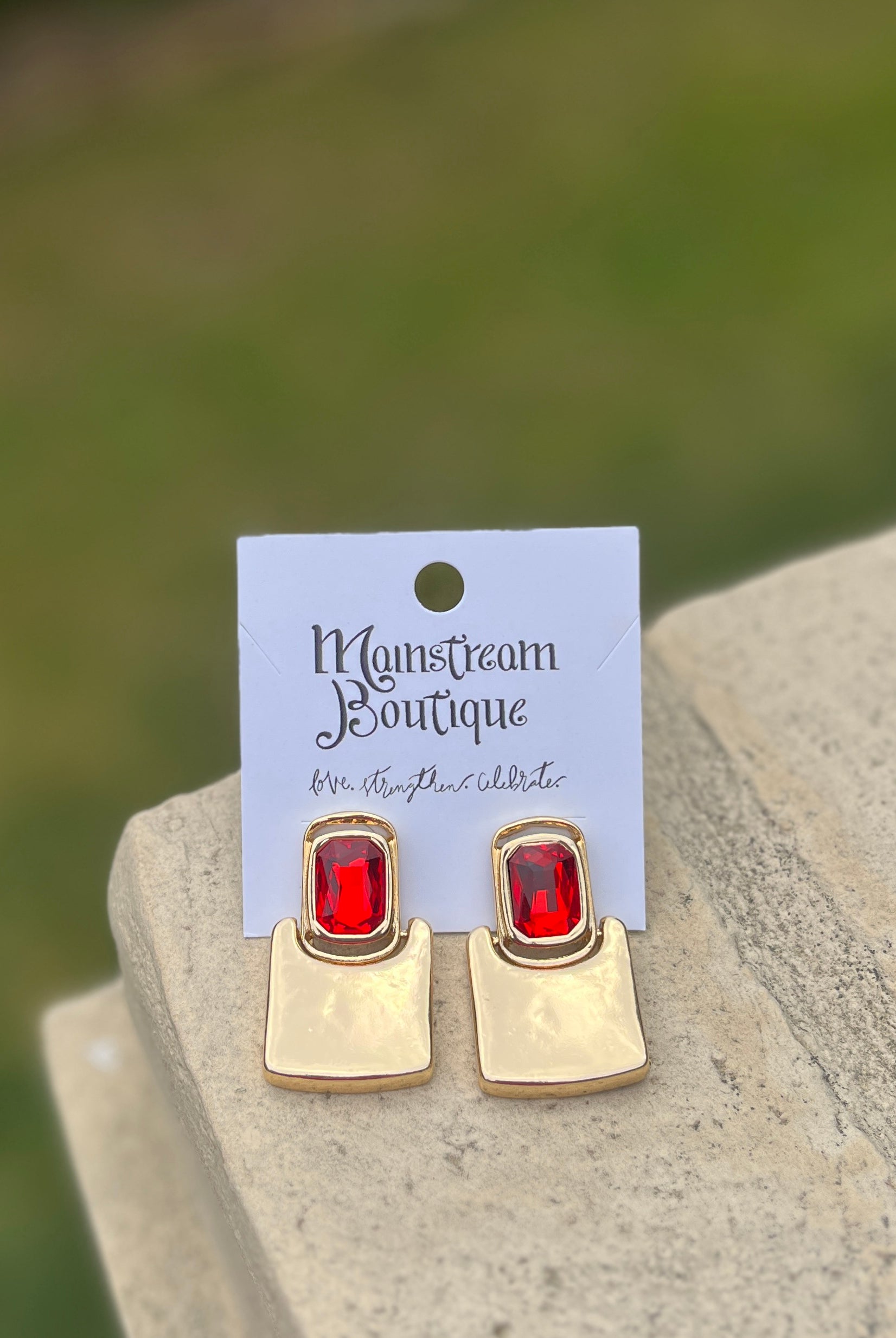 Gold earrings with red stones against a blurred green background.