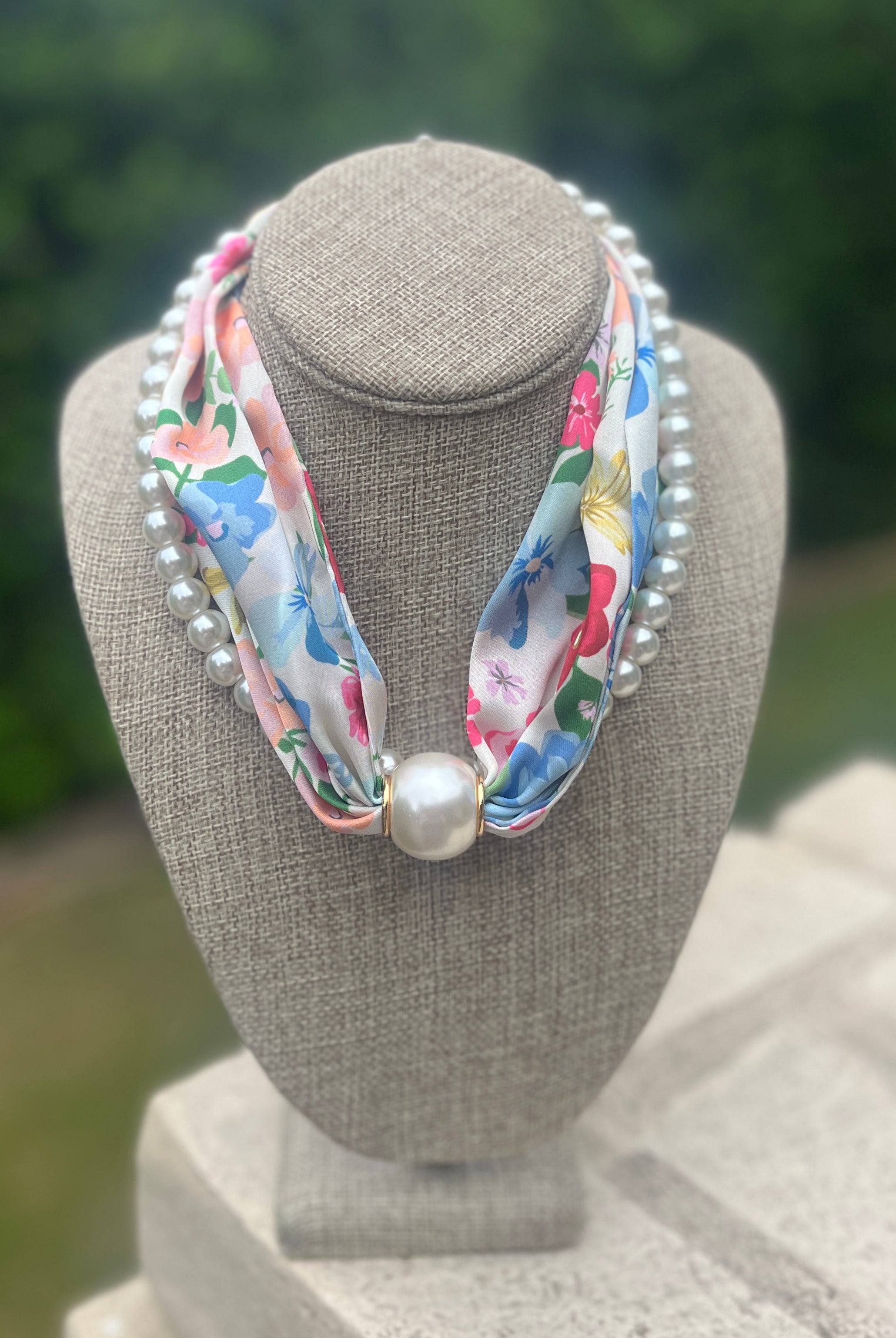 Necklace scarf with floral ribbon and pearls on a gray bust against a blurred green background