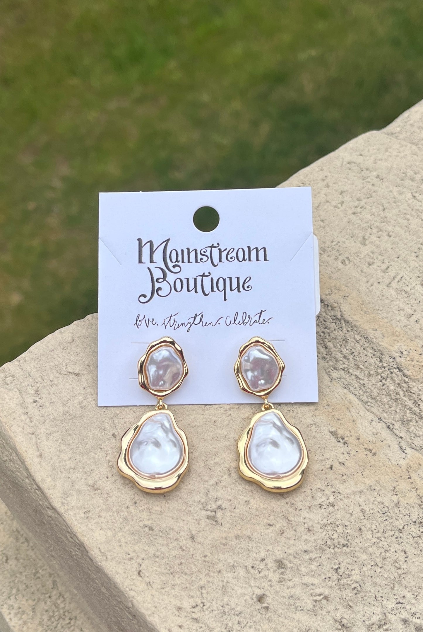 Pair of gold and Mother of Pearl teardrop earrings against a stone and grass background.