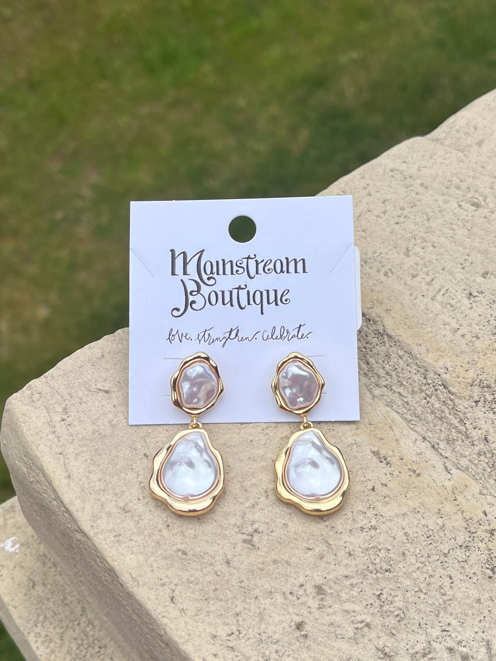 Pair of gold and Mother of Pearl teardrop earrings against a stone and grass background.