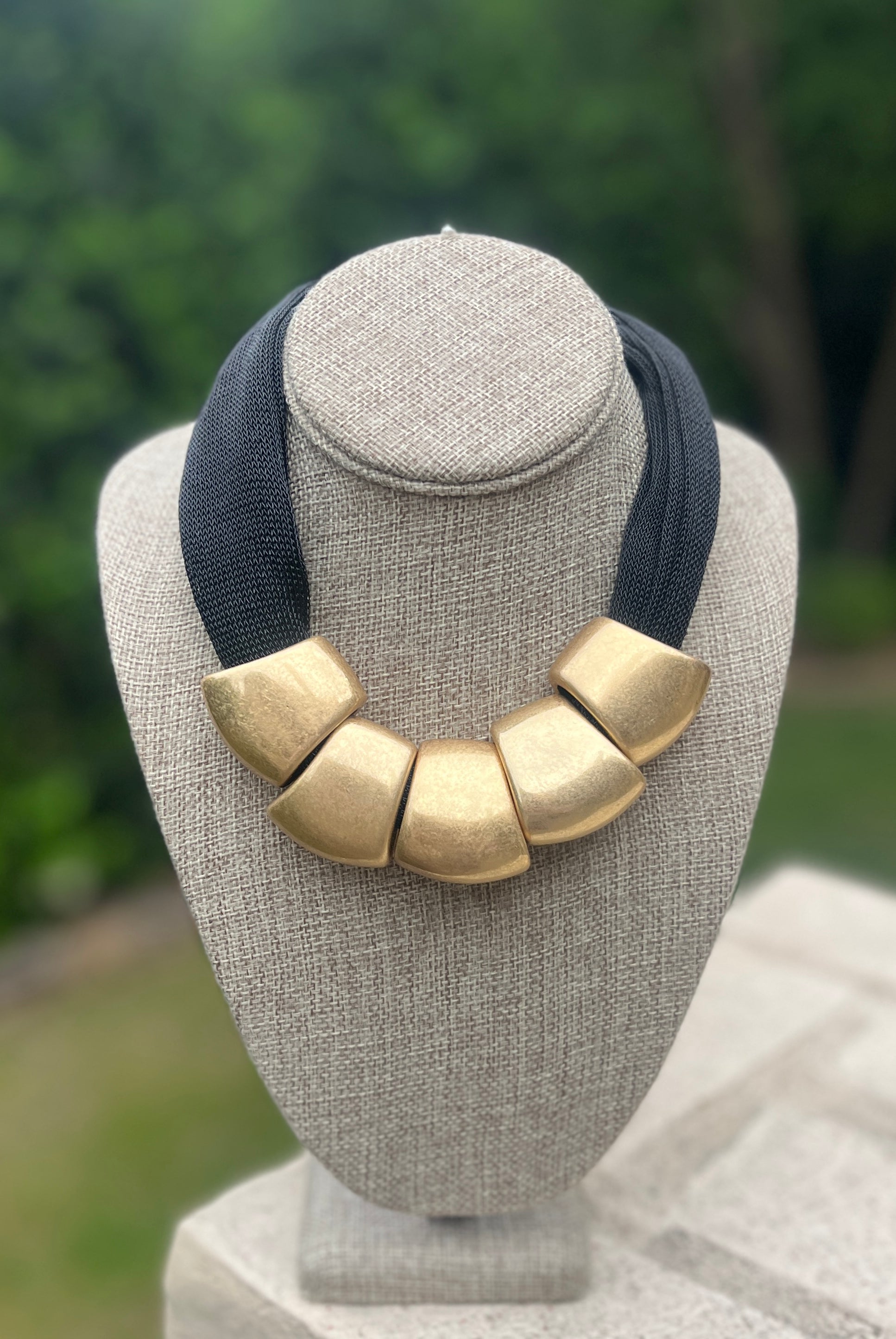 Necklace with gold bibs and navy strap on a gray mannequin bust with a blurred green background