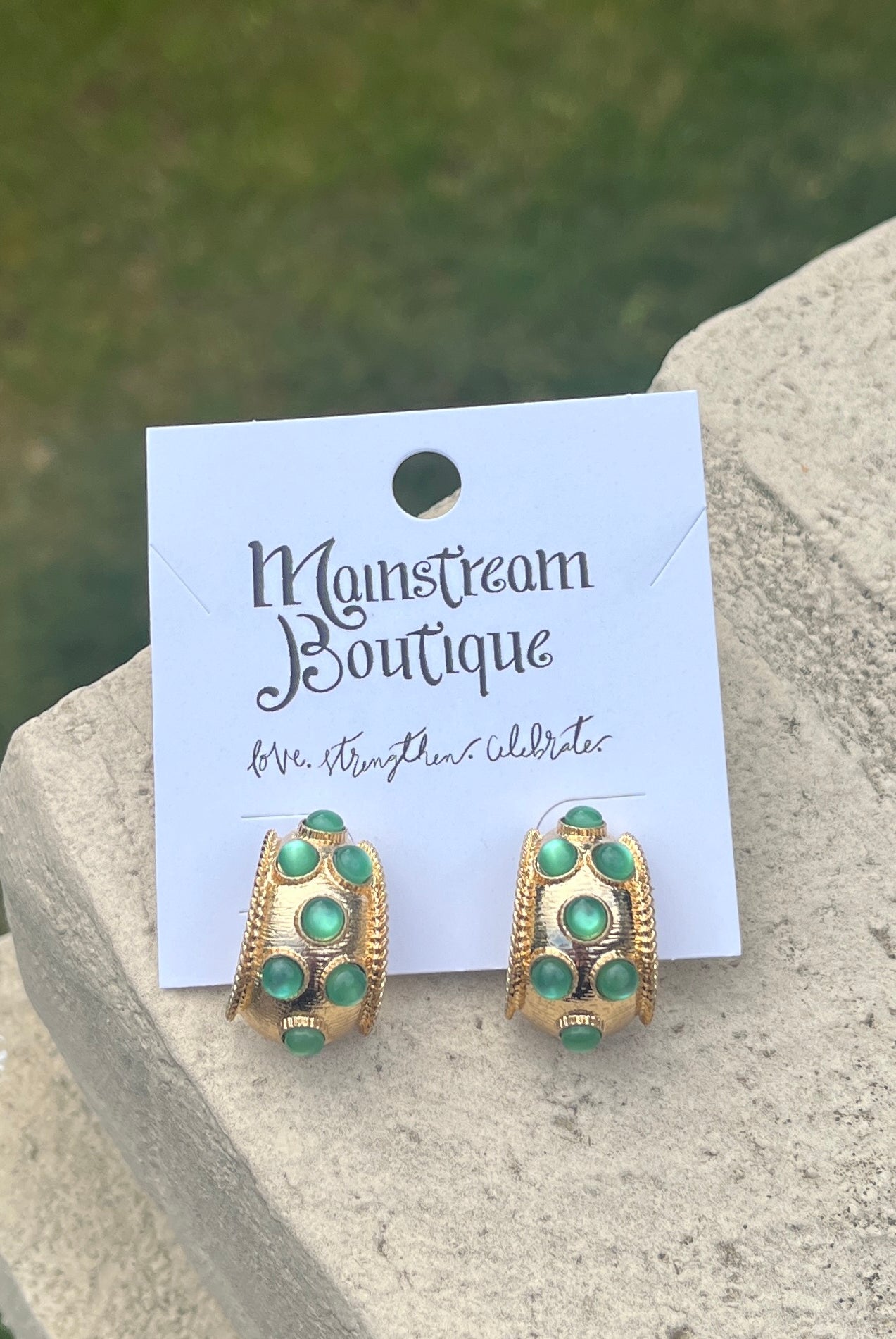 Gold and green earrings against a stone and grass background