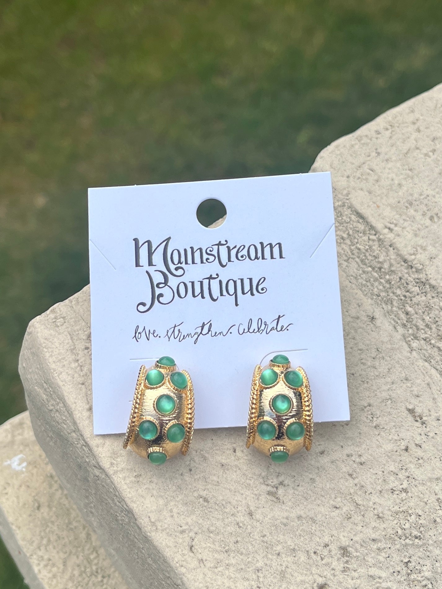 Gold and green earrings against a stone and grass background