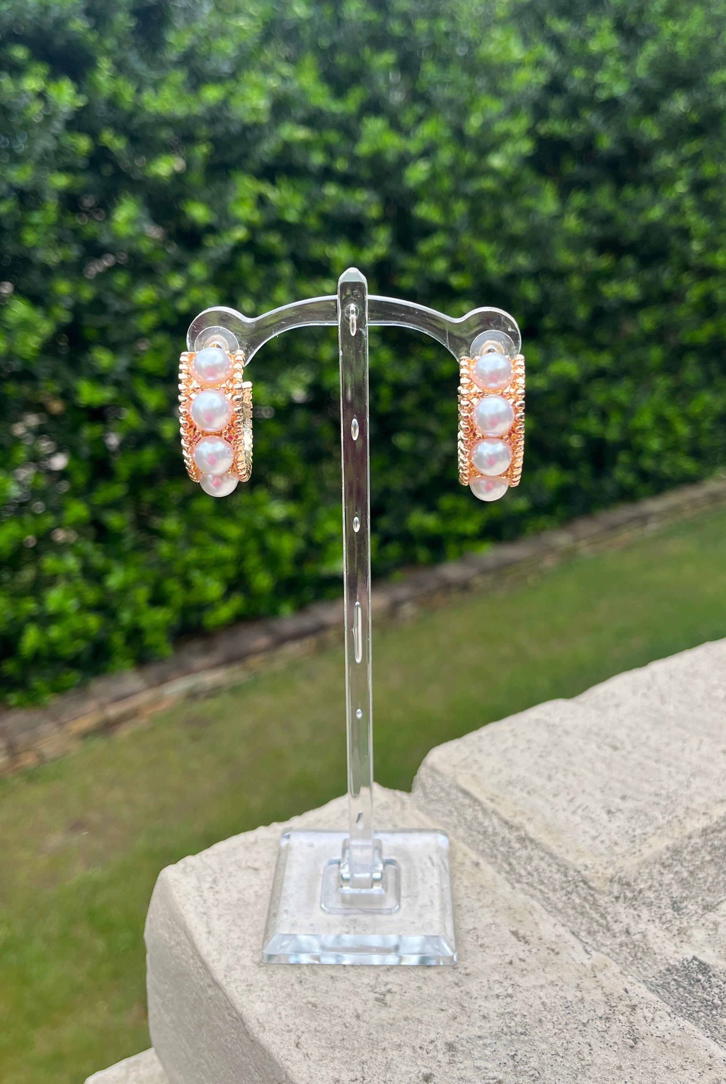Pair of gold/pearl earrings on a clear stand with a green outdoor background