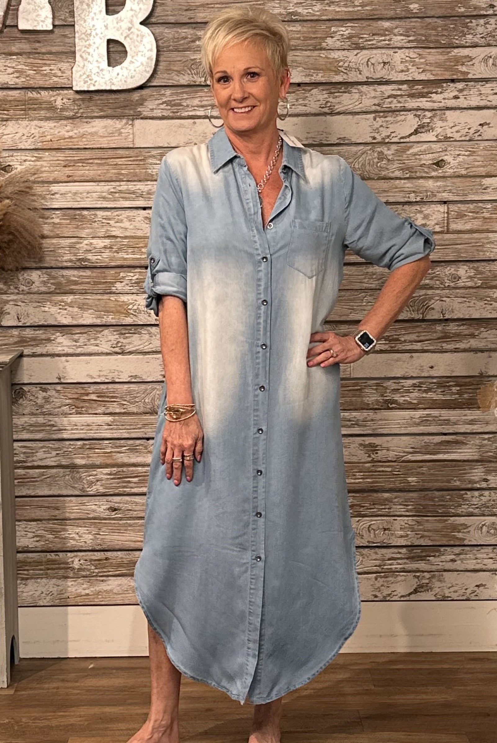 A woman modeling a light blue denim button-down shirt dress with a faded white wash effect, standing in front of a rustic wood-paneled wall.