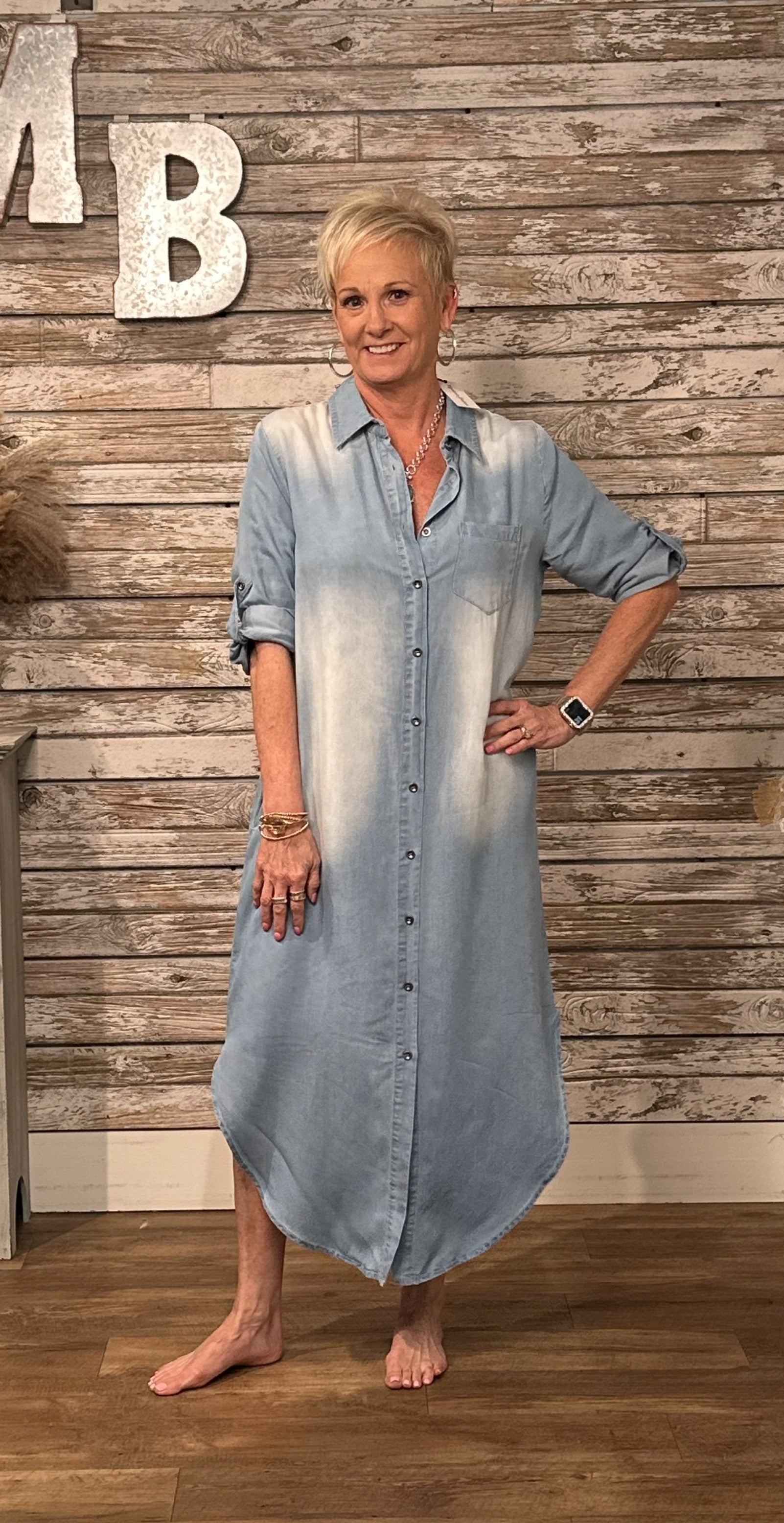 A woman modeling a light blue denim button-down shirt dress with a faded white wash effect, standing in front of a rustic wood-paneled wall.