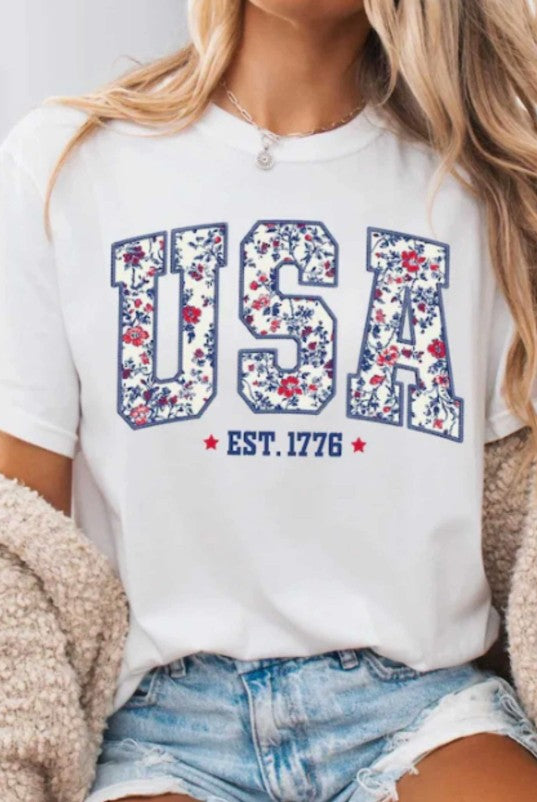 Person wearing a white t-shirt with 'USA' and floral design, sitting on a couch.