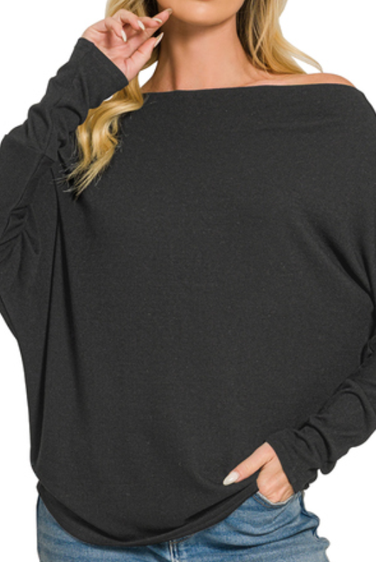 A woman wearing a black off-the-shoulder long-sleeve knit top with a relaxed drape, paired with blue jeans, against a plain white background.
