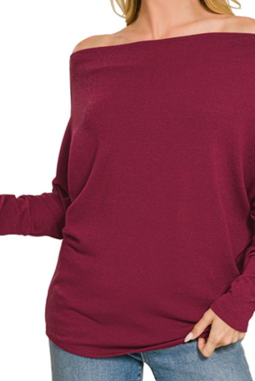 A woman wearing a burgundy off-the-shoulder long-sleeve knit top with gathered sleeves, paired with blue jeans, shown against a plain white background.