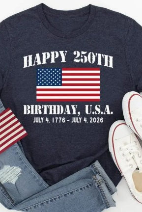 Navy t-shirt with American flag design celebrating the 250th birthday of the USA, paired with denim jeans and white sneakers on a light gray background.