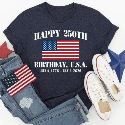 Navy t-shirt with American flag design celebrating the 250th birthday of the USA, paired with denim jeans and white sneakers on a light gray background.