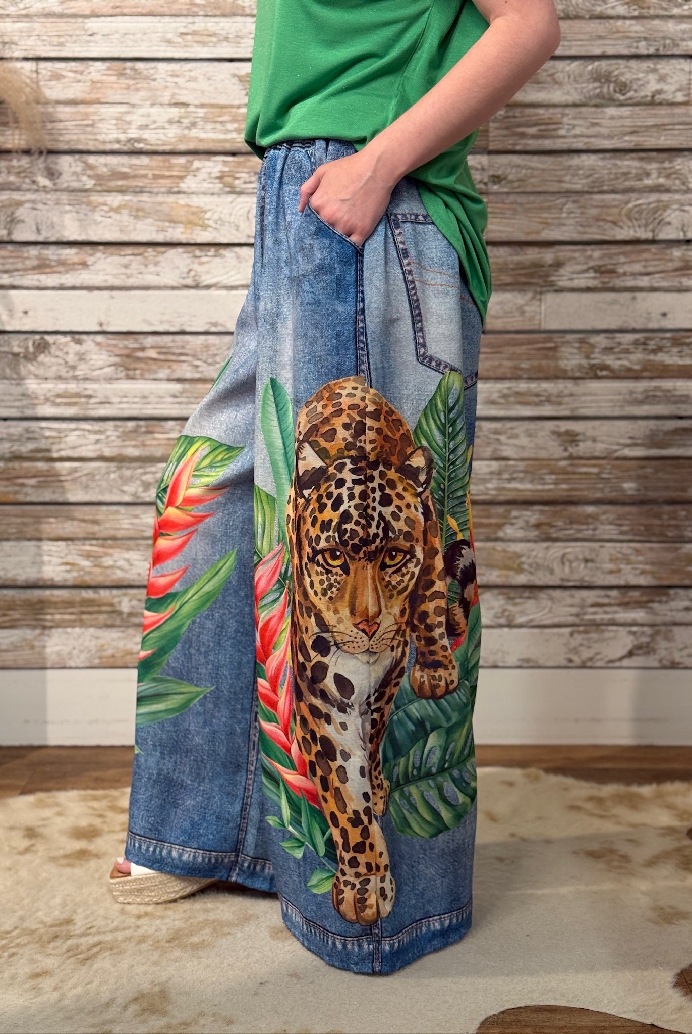 Person wearing a pair of wide-leg jeans with a leopard and tropical leaf design against a wooden wall.