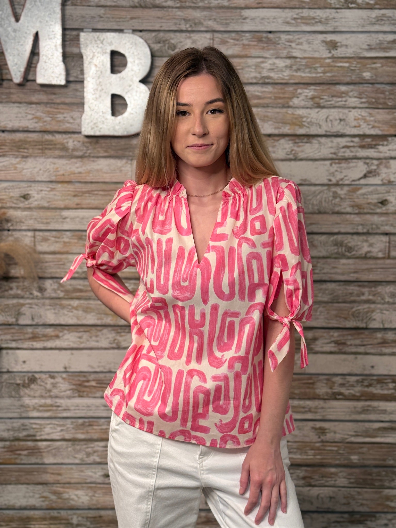 Woman wearing a pink patterned blouse standing against a wooden wall with decorative letters.