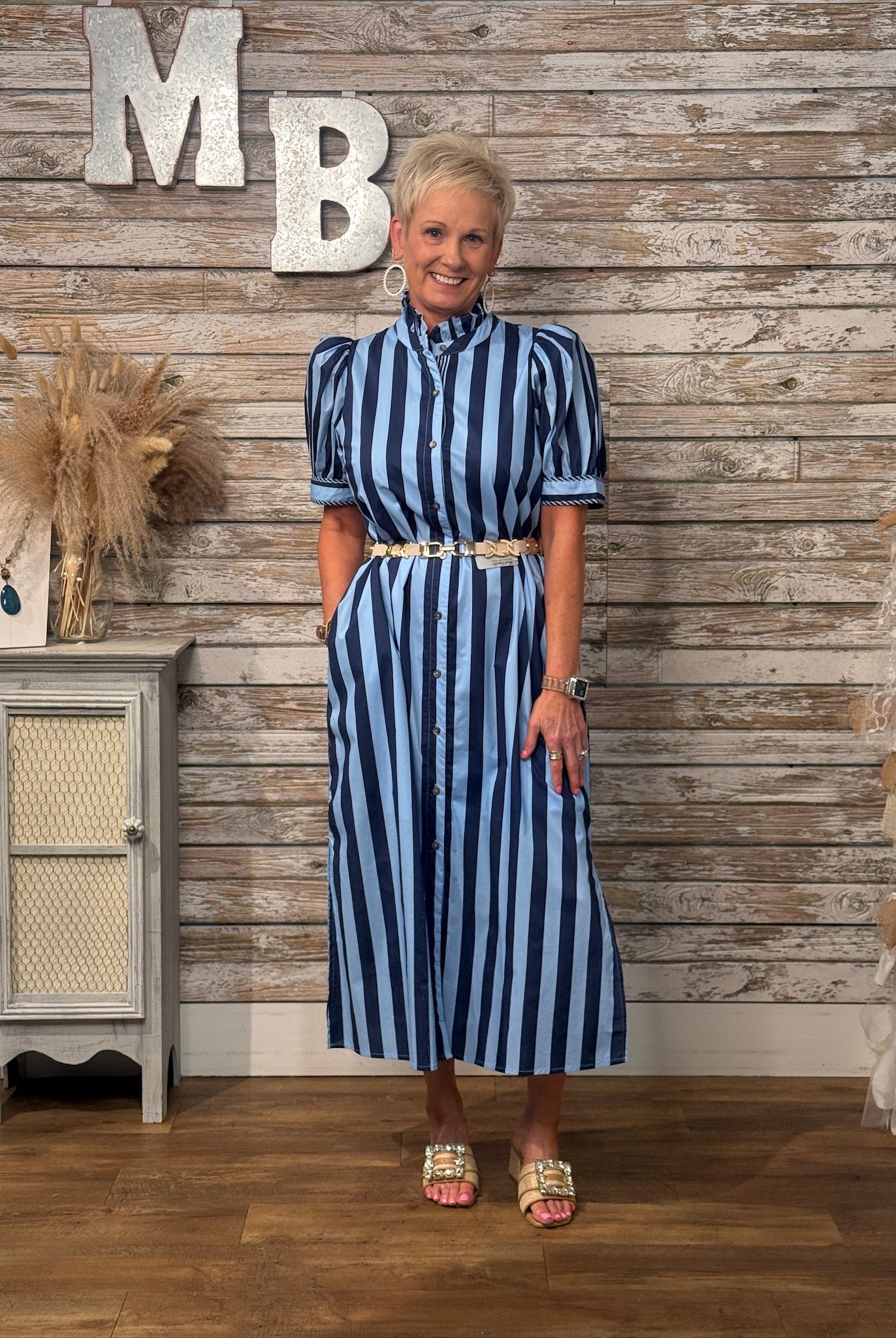 A woman modeling a navy and light blue vertically striped midi dress with puff sleeves and a gold belt, standing in front of a rustic wood-paneled wall with decorative letters and a small cabinet.