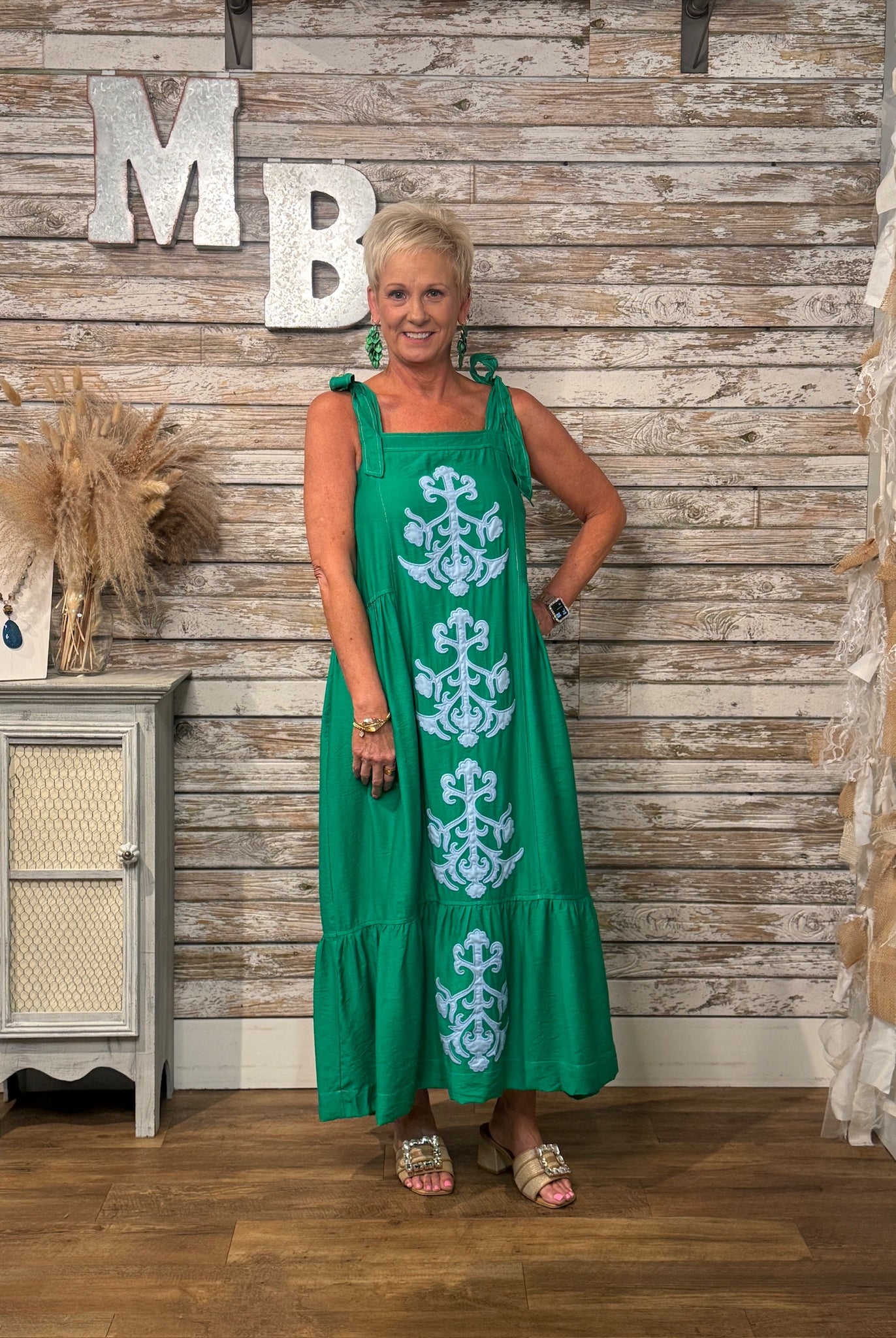 A woman modeling a green sleeveless maxi dress with light blue anchor motifs, standing in front of a rustic wood-paneled wall with decorative letters and a small cabinet.