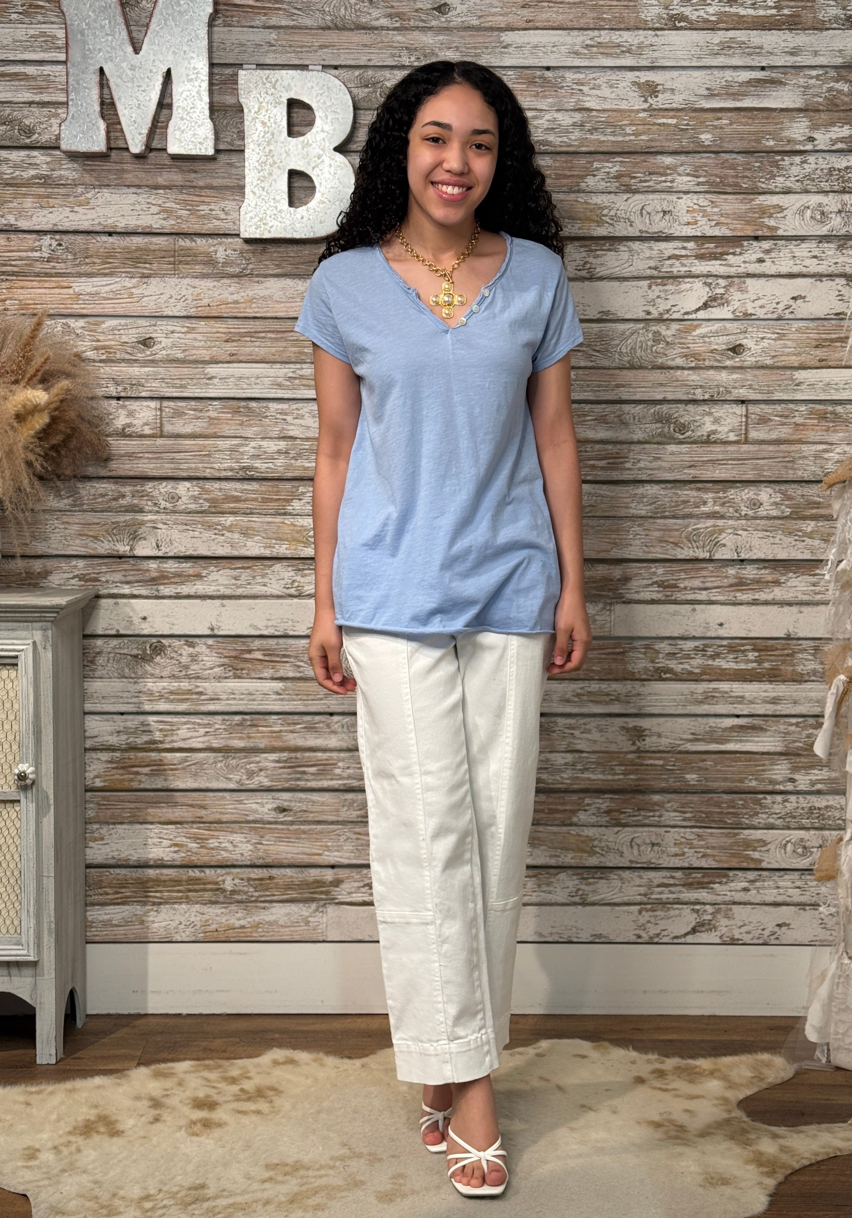 Woman wearing a blue top and white pants standing against a wooden wall with decorative letters.