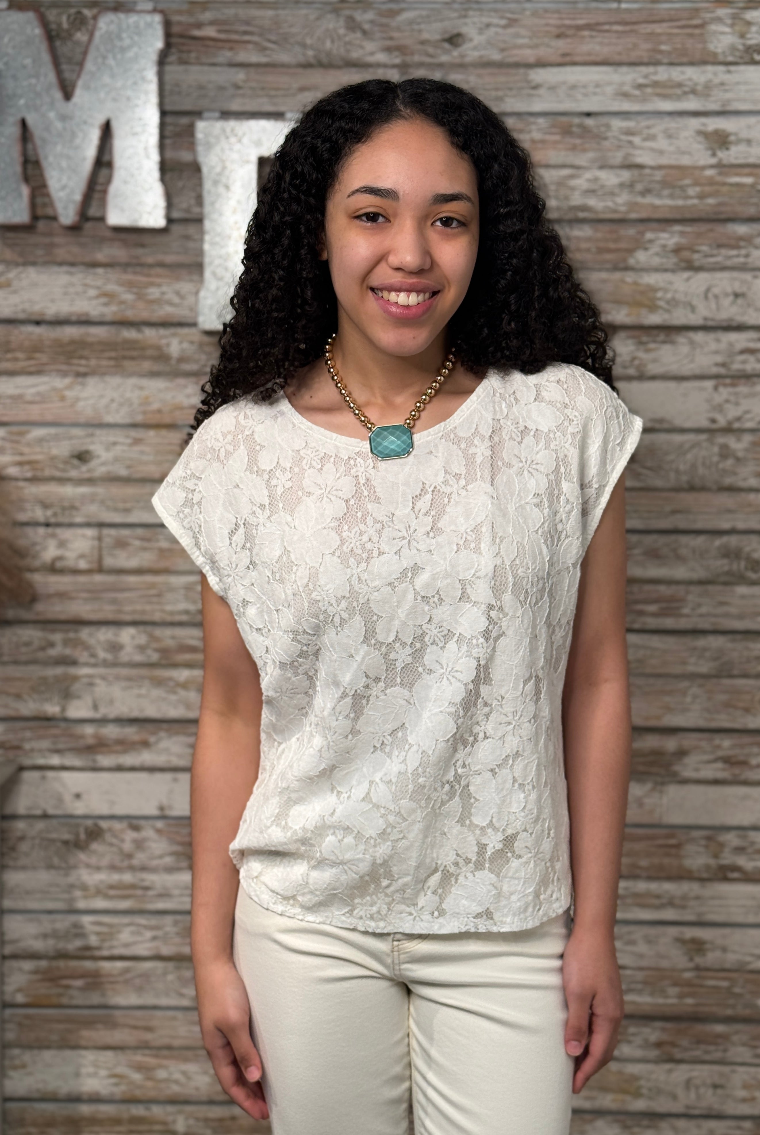 Woman wearing a white lace top and beige pants against a wooden wall with decorative letters.
