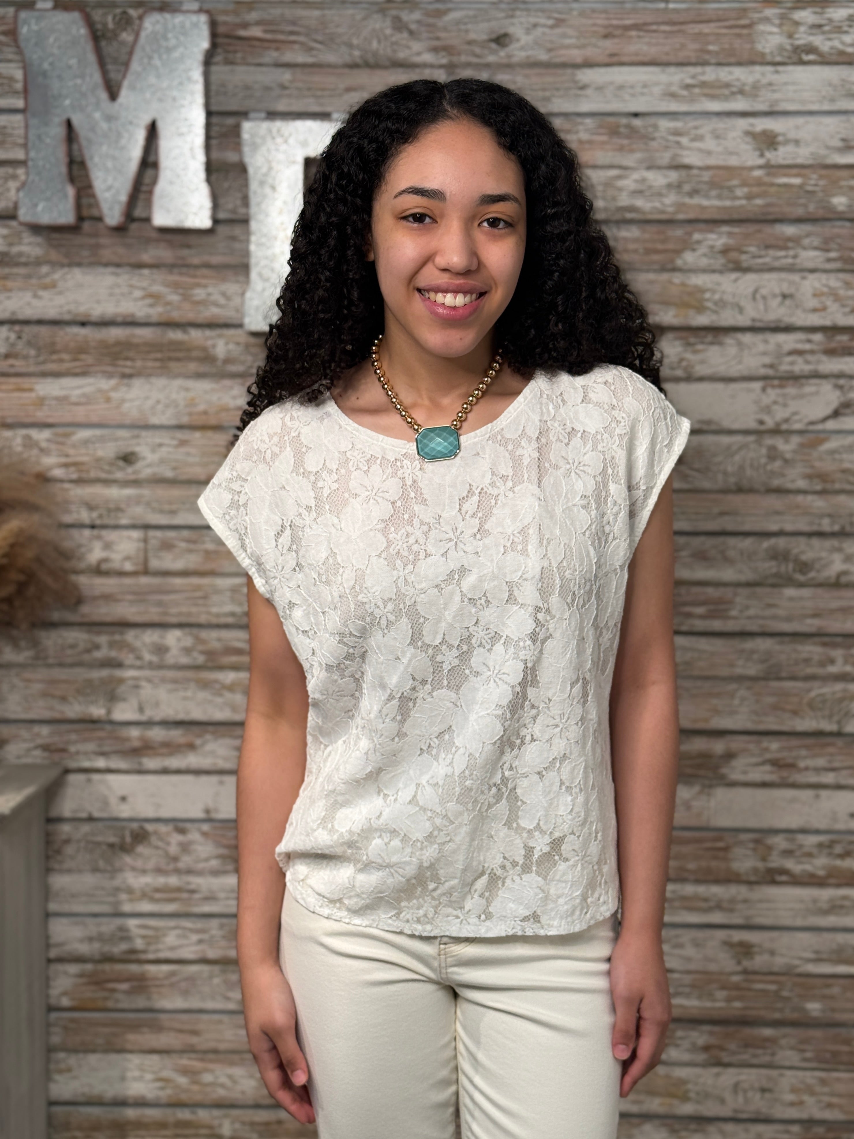 Woman wearing a white lace top and beige pants against a wooden wall with decorative letters.