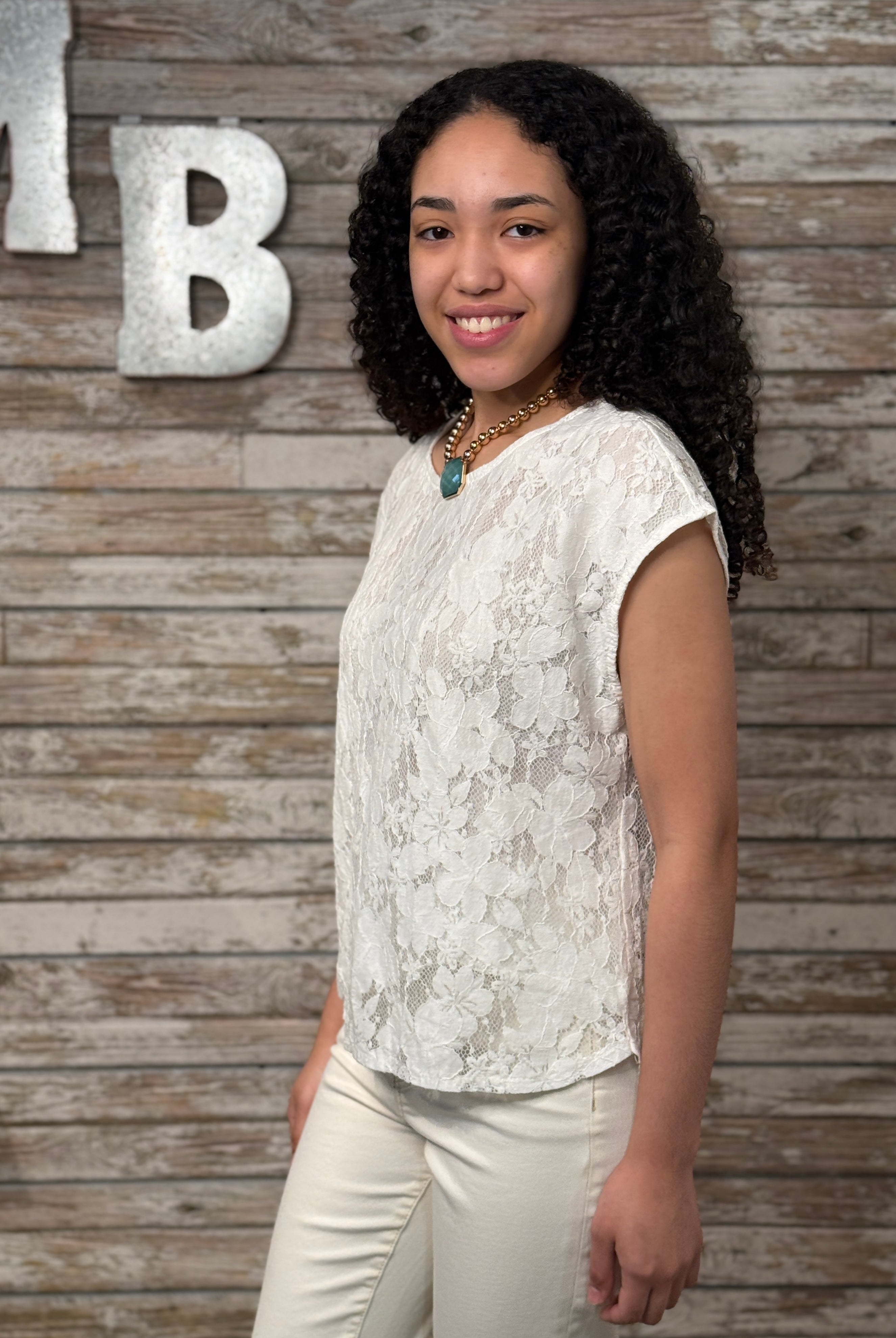 Woman in a white lace top and beige pants standing against a wooden wall with decorative letters.