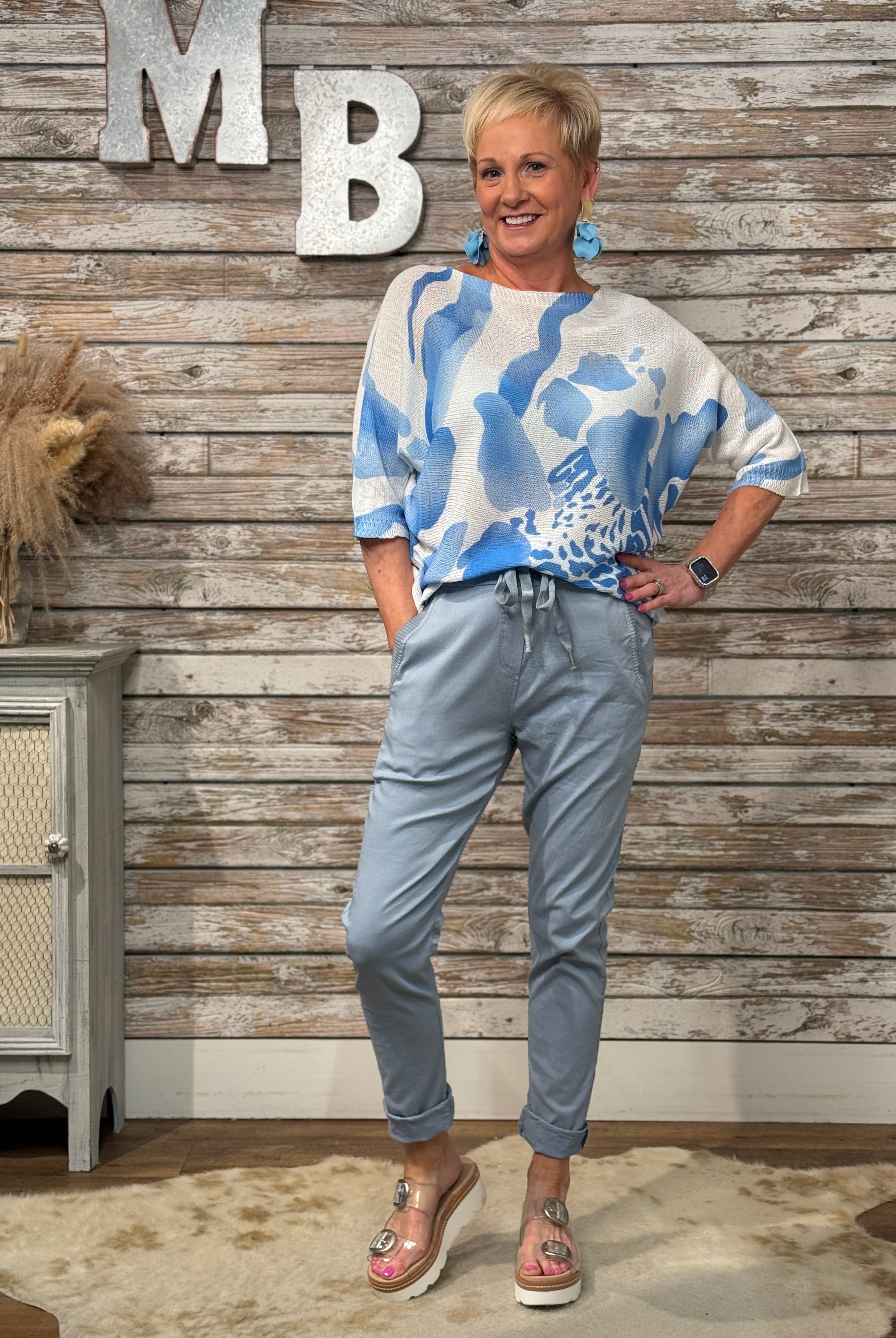Woman wearing a blue and white patterned sweater and light blue jeans standing against a wooden wall.