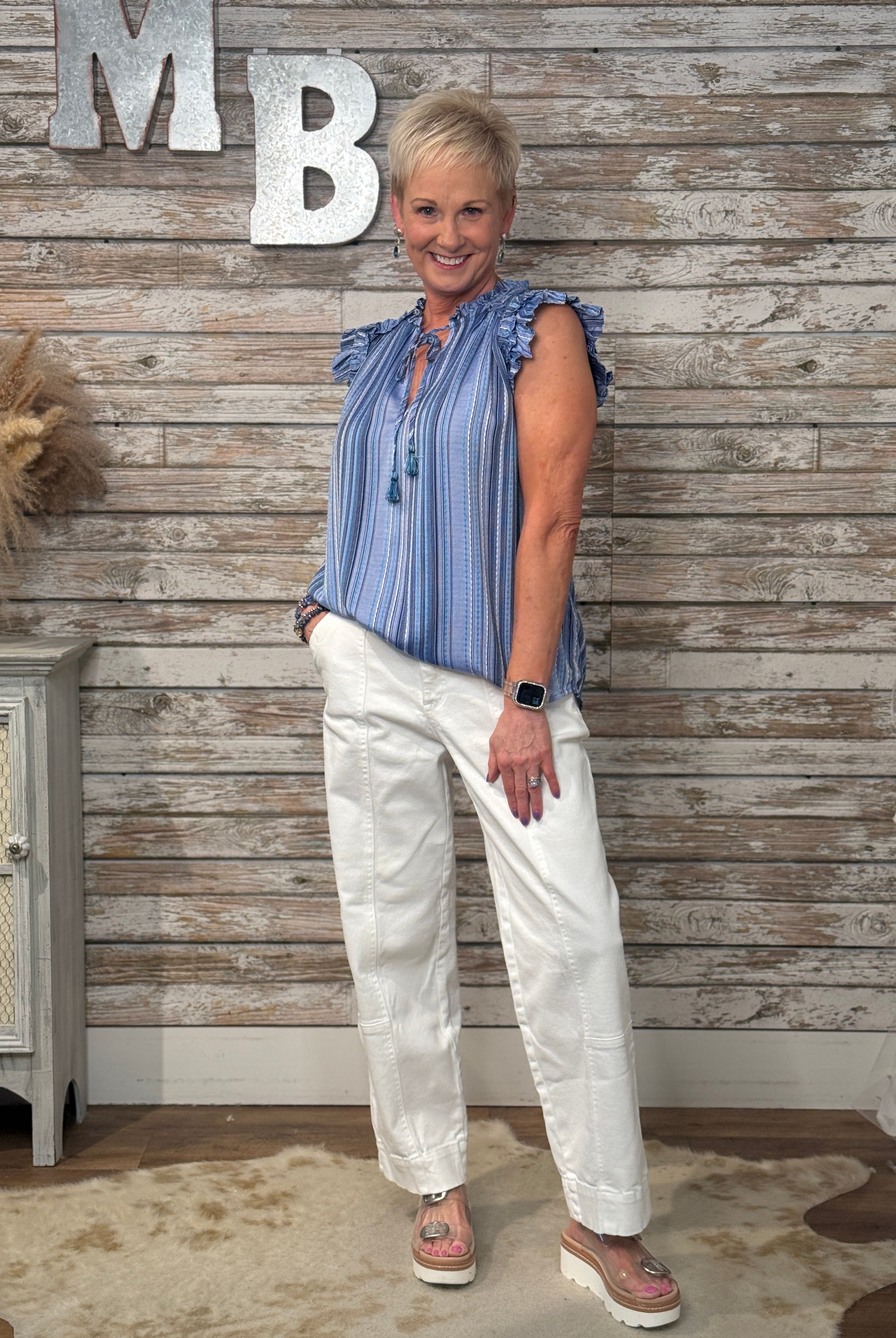 Woman in a blue striped shirt and white pants standing against a wooden wall with decorative letters.
