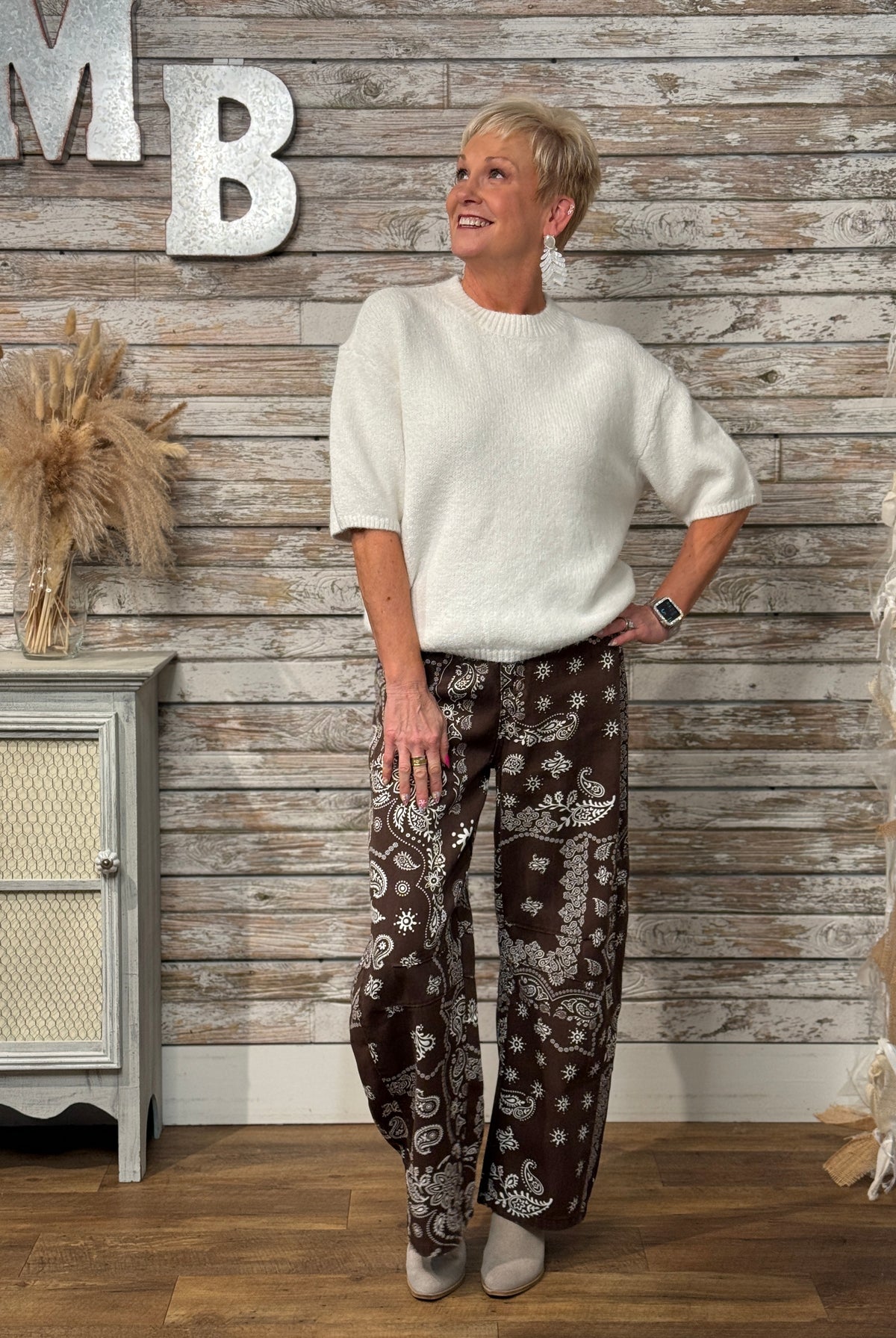 A woman stands in a rustic indoor setting wearing a cream short-sleeve knit top and dark brown wide-leg pants with a white paisley pattern. She is posed in front of a weathered wood plank wall with decorative silver letters and a small cabinet to the side.