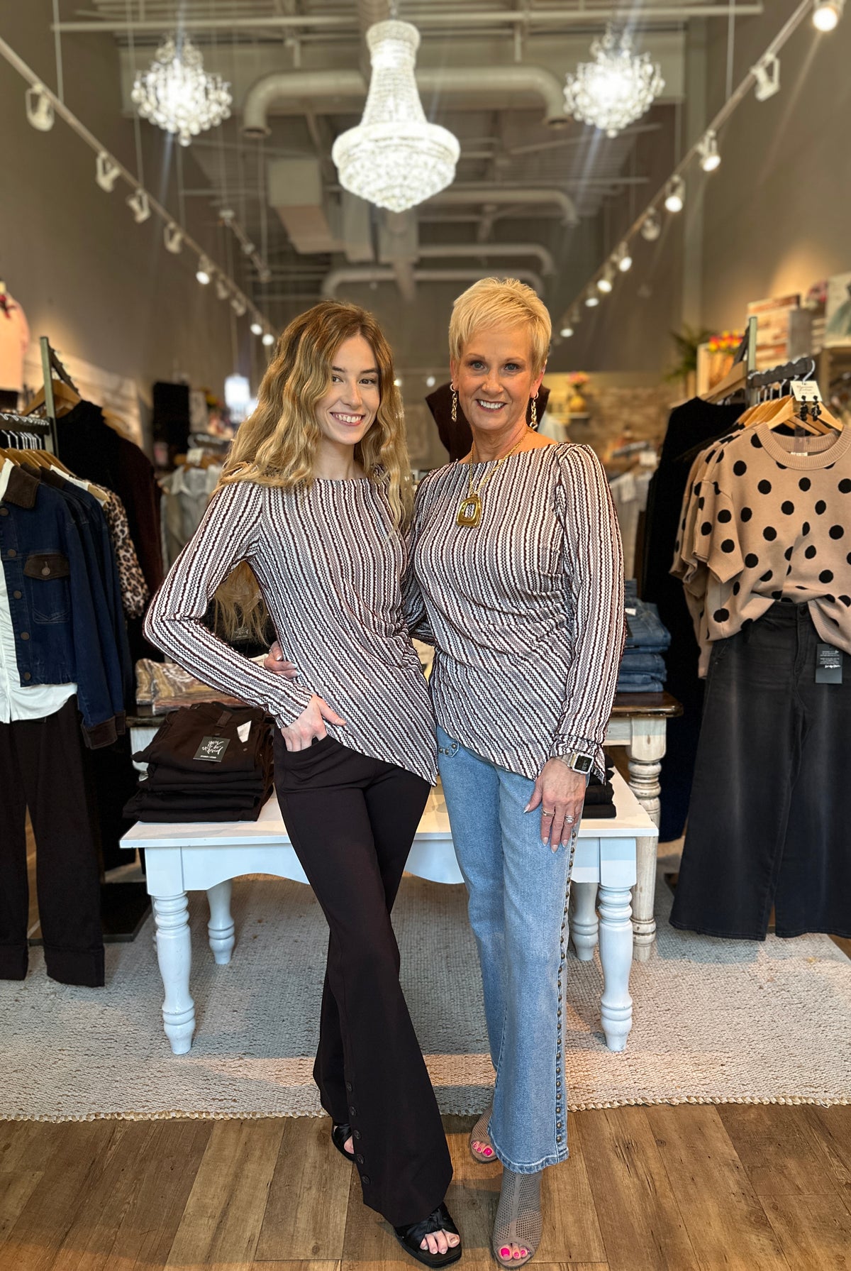Two women posing in a boutique store, both wearing matching striped long-sleeve top and jeans. The woman on the left wears a black and white striped top with black pants, while the woman on the right wears a black and white striped top with light blue jeans.
