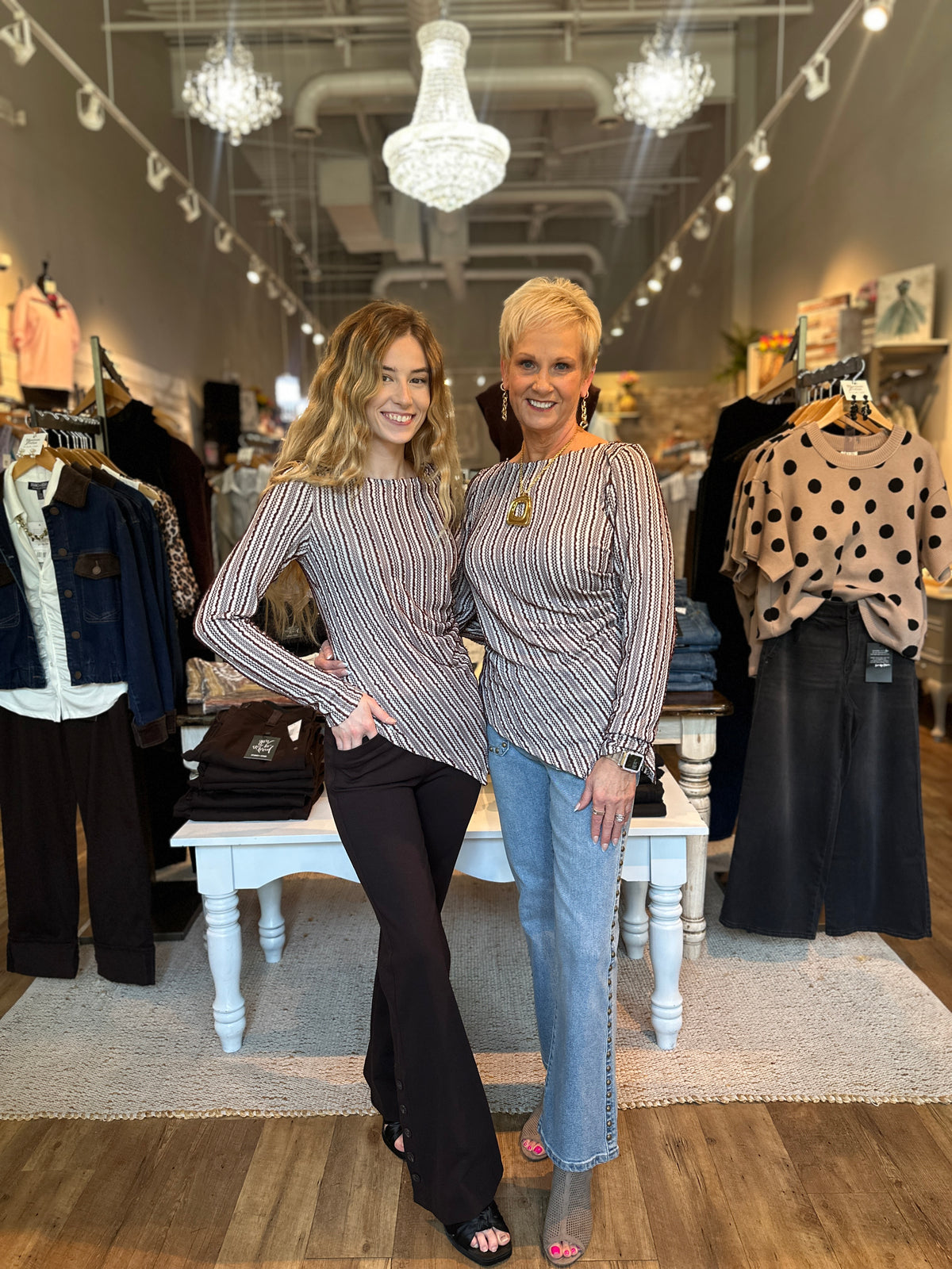 Two women posing in a boutique store, both wearing matching striped long-sleeve top and jeans. The woman on the left wears a black and white striped top with black pants, while the woman on the right wears a black and white striped top with light blue jeans.