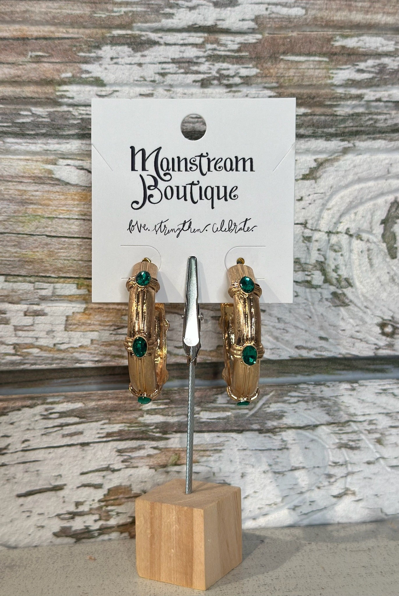 Gold hoop earrings with green stones on a wooden stand against a rustic wood background.