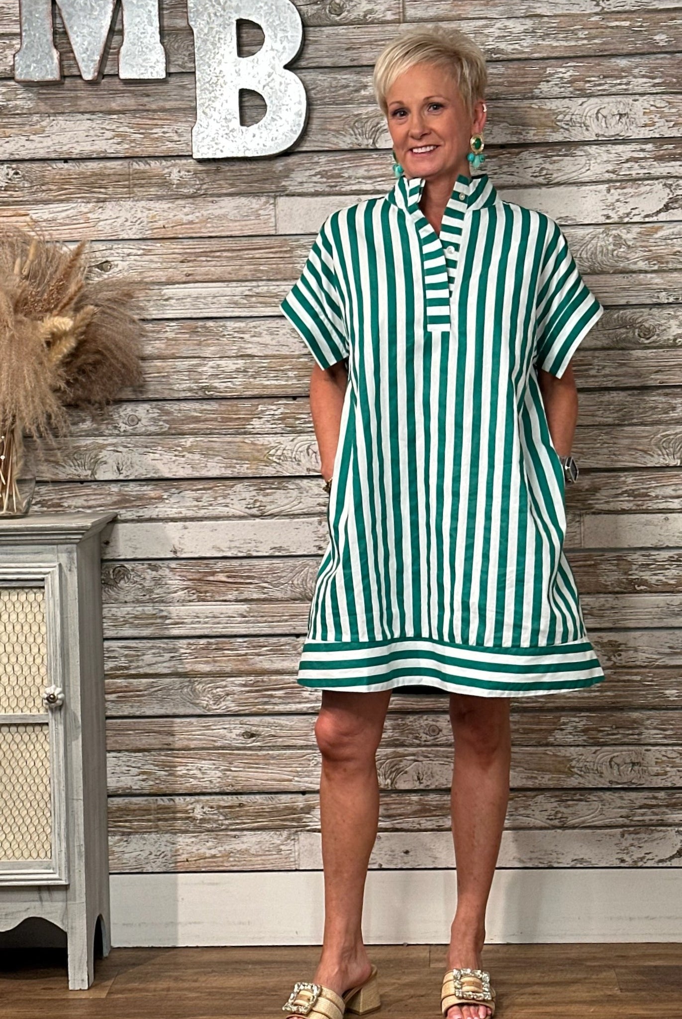 A woman modeling a green and white vertically striped short-sleeve tunic dress with a high collar and front pockets, standing in front of a rustic wood-paneled wall with decorative letters.