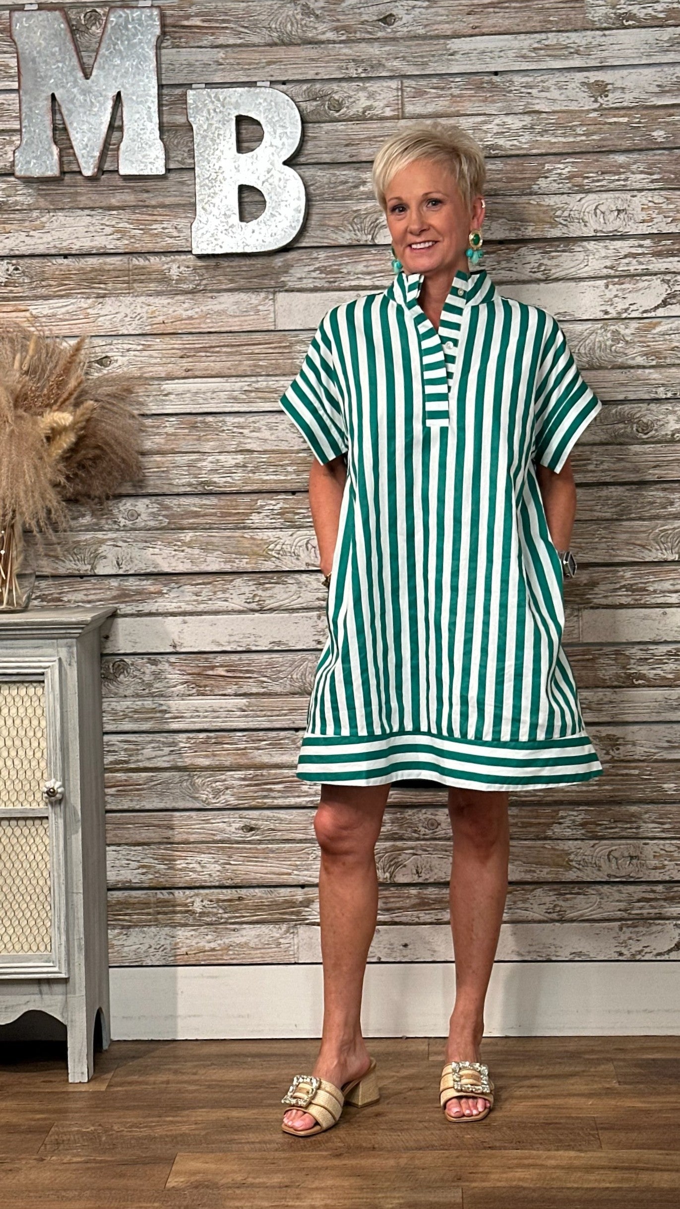 A woman modeling a green and white vertically striped short-sleeve tunic dress with a high collar and front pockets, standing in front of a rustic wood-paneled wall with decorative letters.