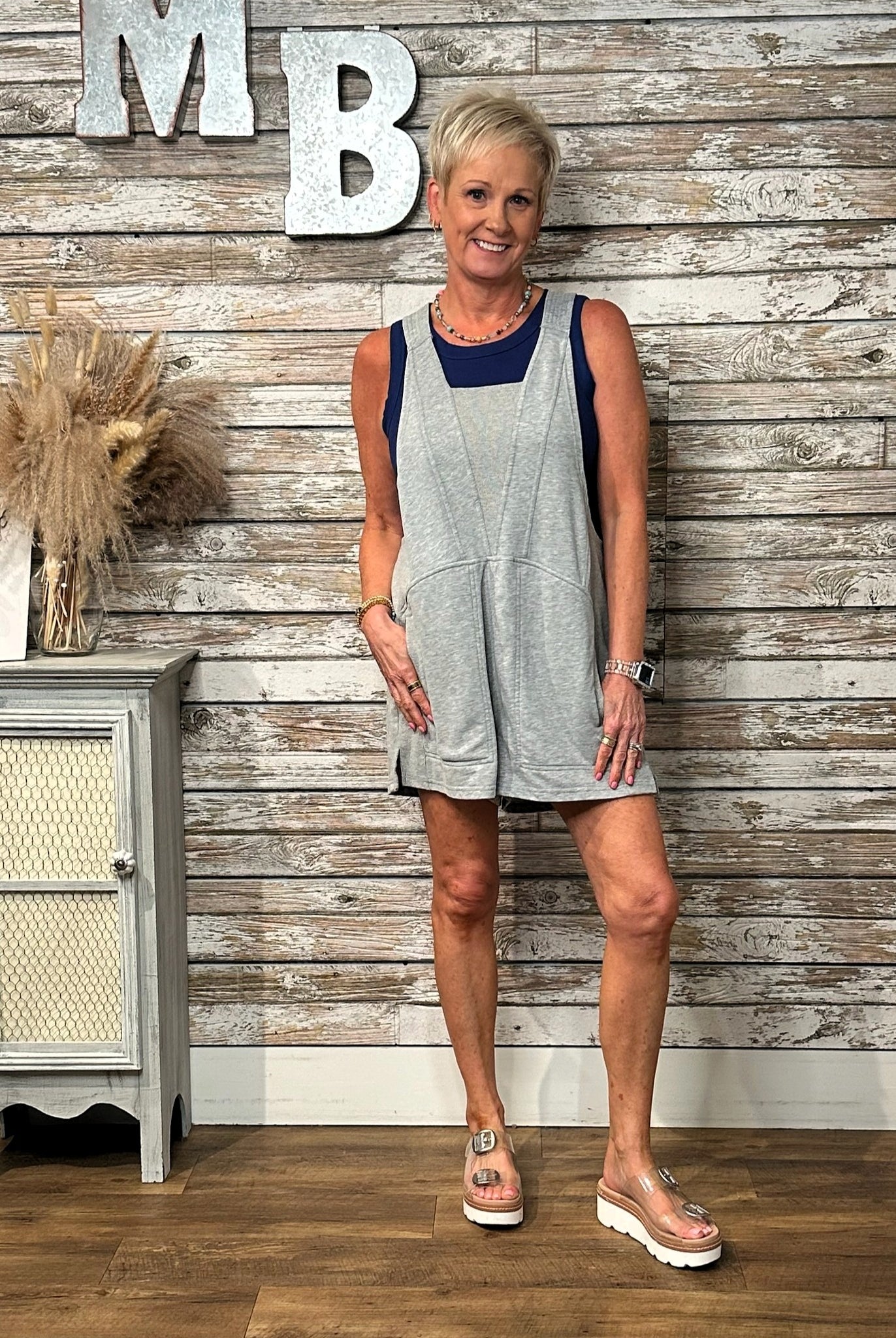 A woman modeling a light gray sleeveless overall romper with a navy blue scoop neckline, standing in front of a rustic whitewashed wood plank wall.