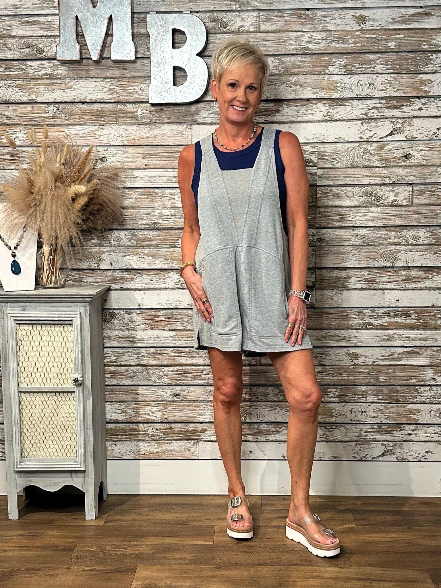 A woman modeling a light gray sleeveless overall romper with a navy blue scoop neckline, standing in front of a rustic whitewashed wood plank wall.