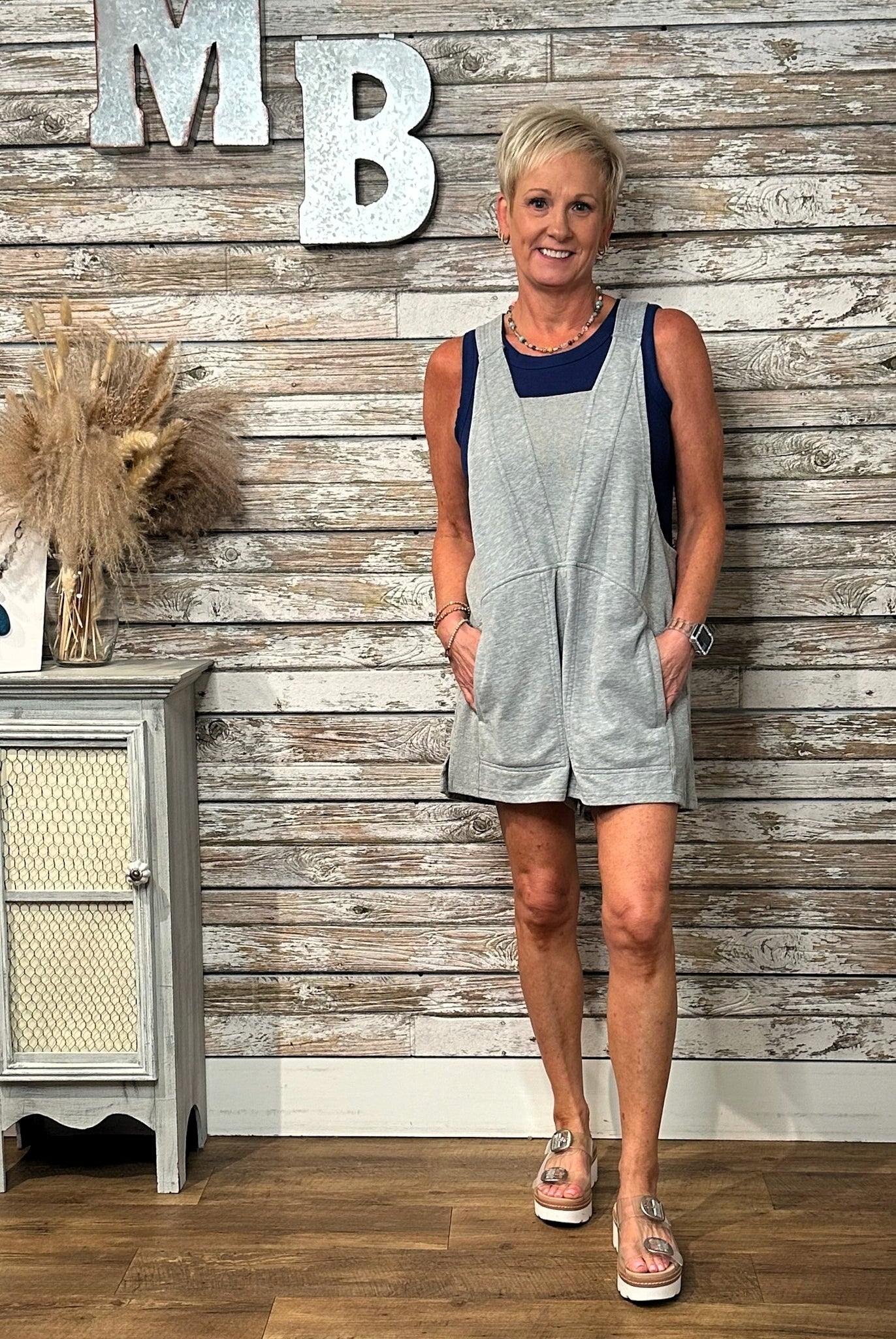 A woman modeling a light gray sleeveless romper with navy blue trim, standing in front of a rustic whitewashed wood plank wall with decorative letters and a small cabinet.