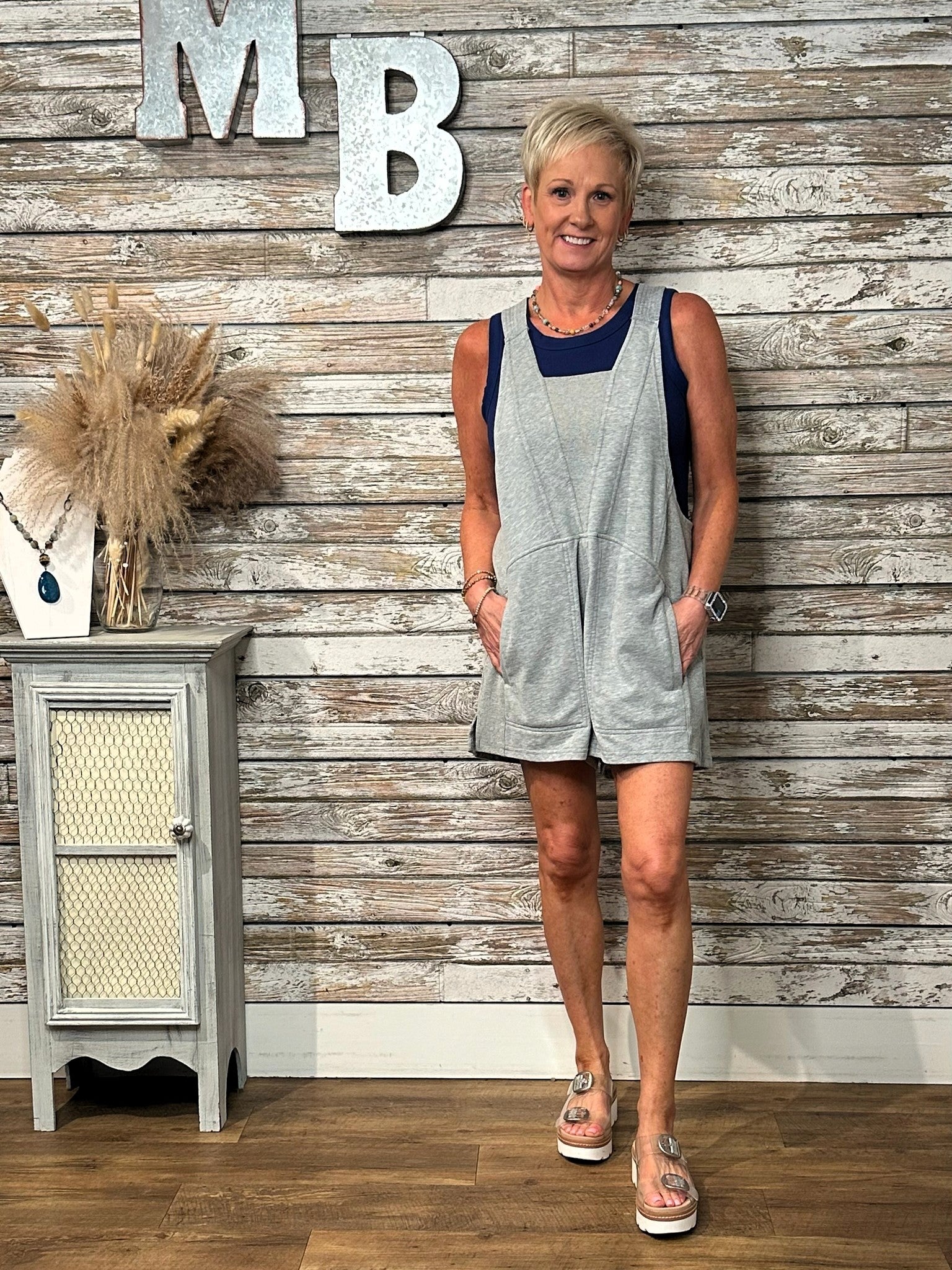 A woman modeling a light gray sleeveless romper with navy blue trim, standing in front of a rustic whitewashed wood plank wall with decorative letters and a small cabinet.