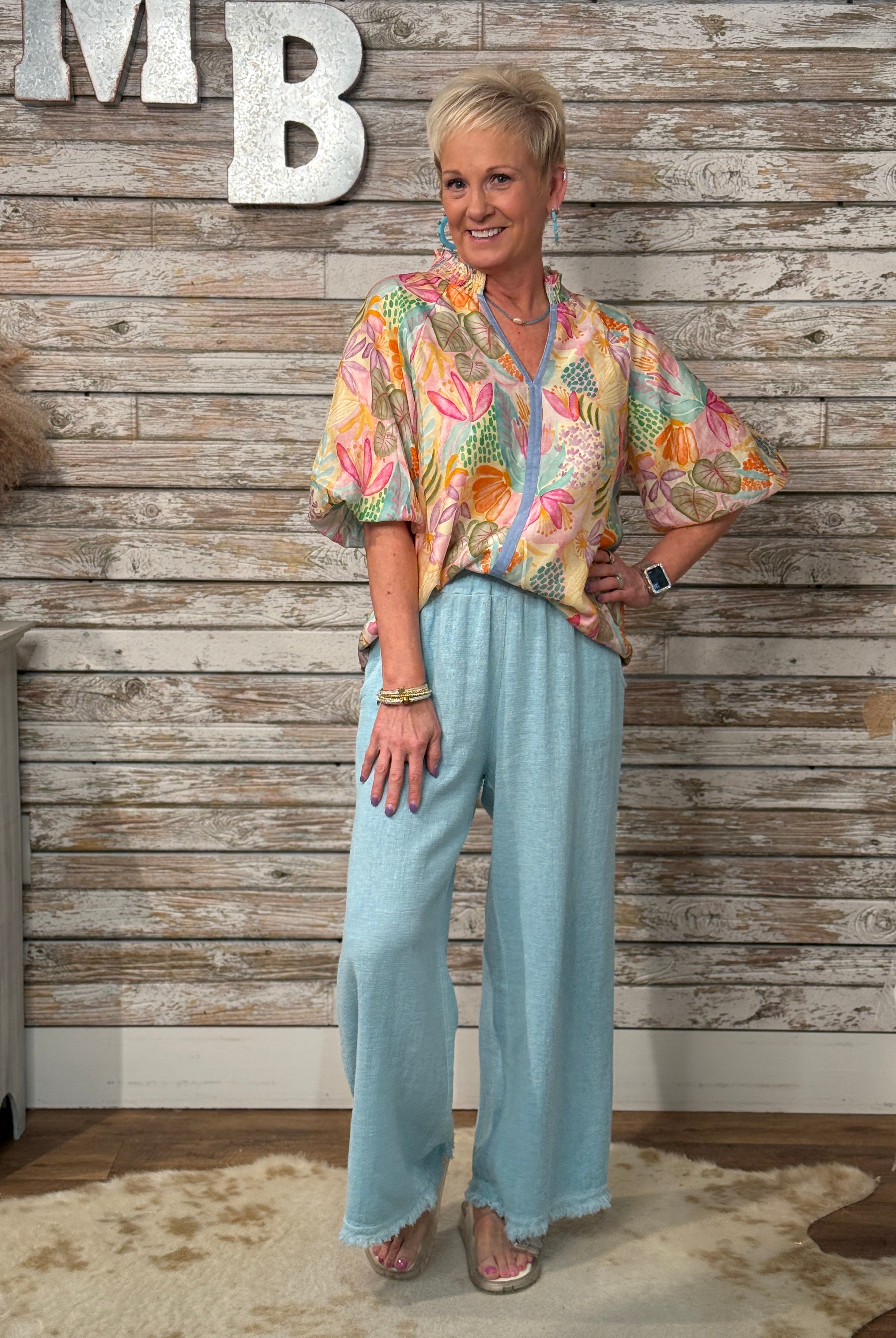 Woman wearing a colorful floral blouse and light blue pants standing against a wooden wall.