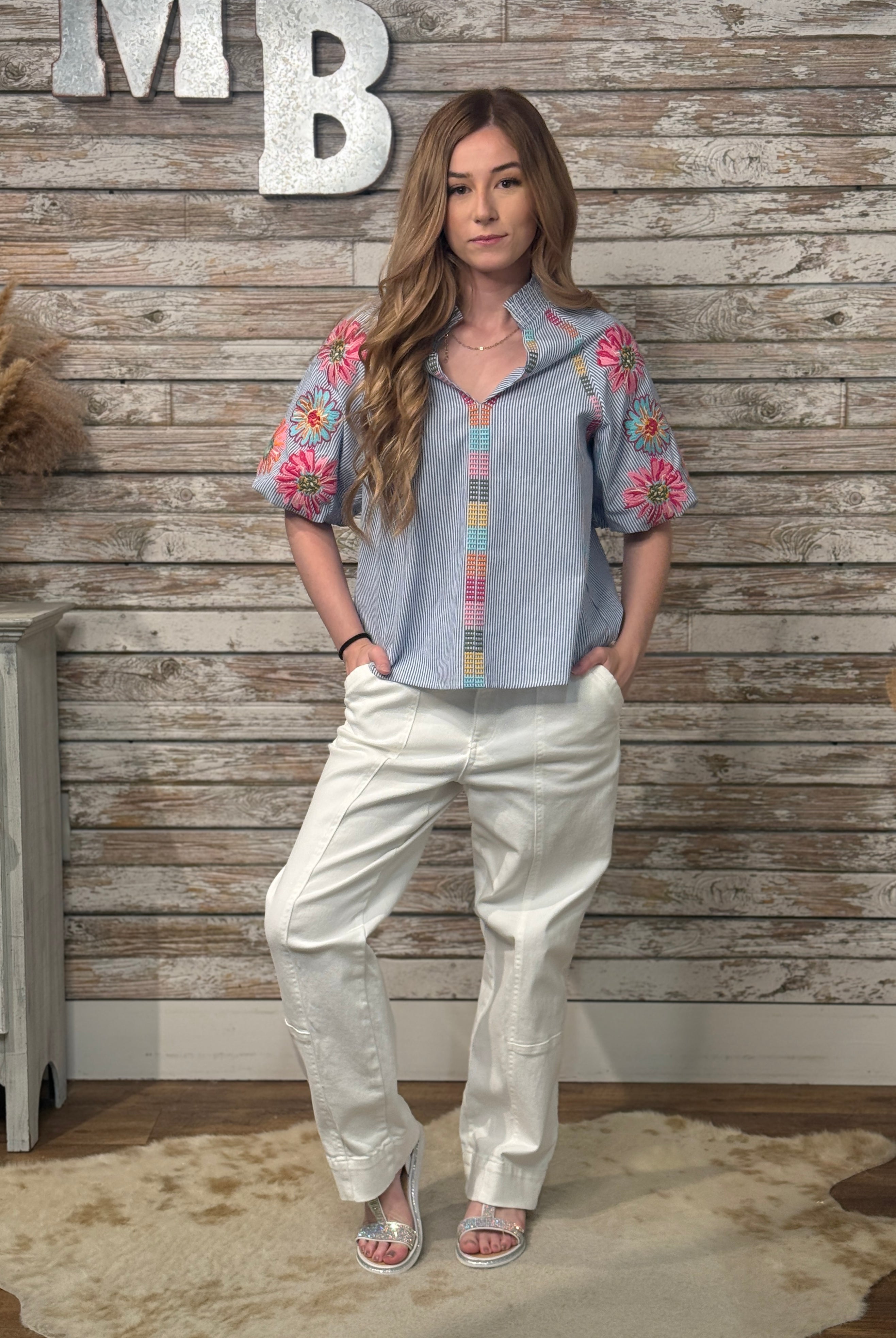 Woman wearing a floral blouse and white pants standing against a wooden wall with decorative elements.
