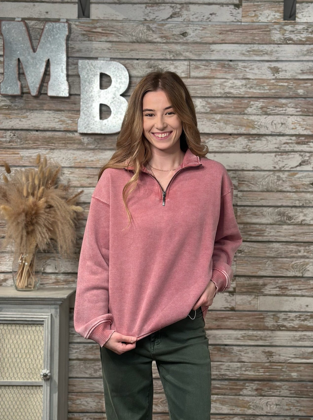 A woman wearing a dusty rose pink quarter-zip fleece pullover sweater and dark green pants, standing in front of a rustic wood-paneled wall with decorative metal letters.