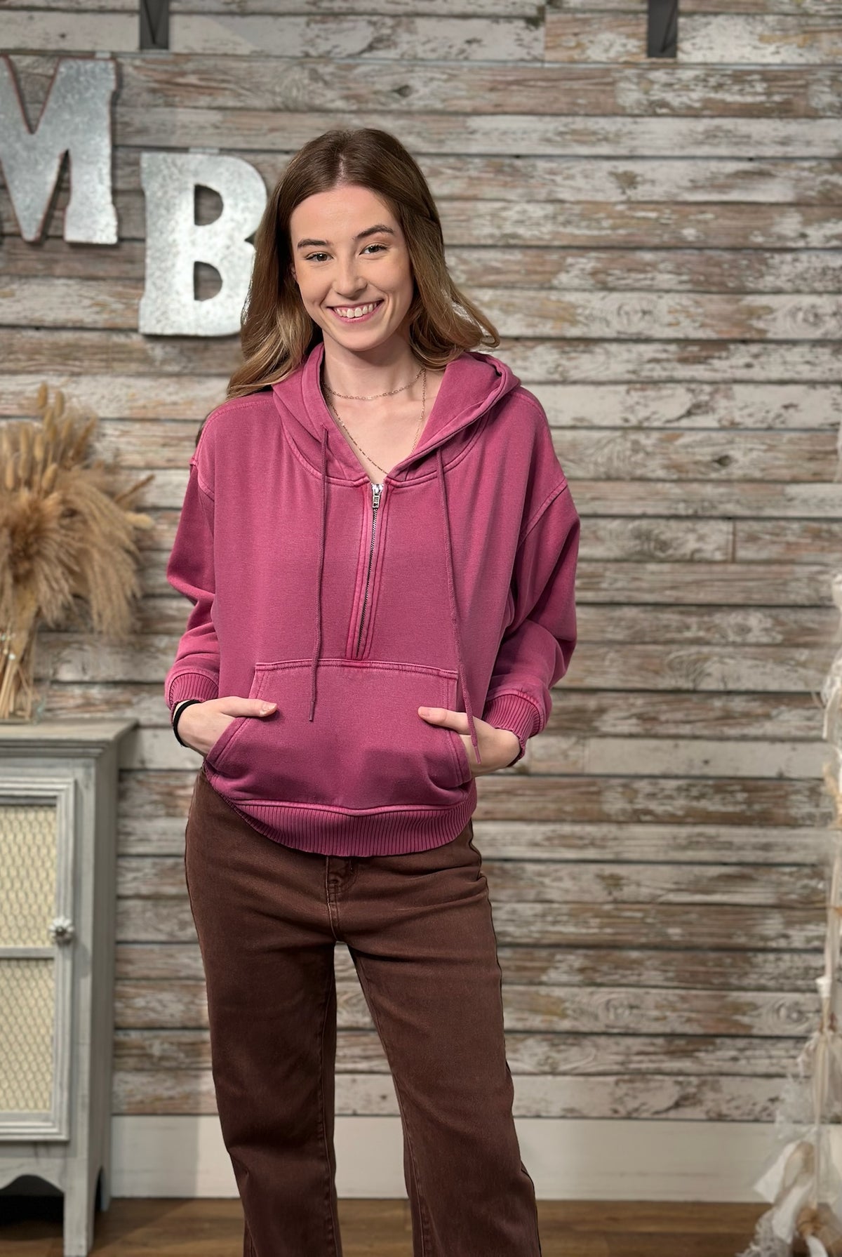 A woman wearing a dusty rose pink half-zip hoodie and brown pants, standing in front of a rustic wood-paneled wall with decorative letters.