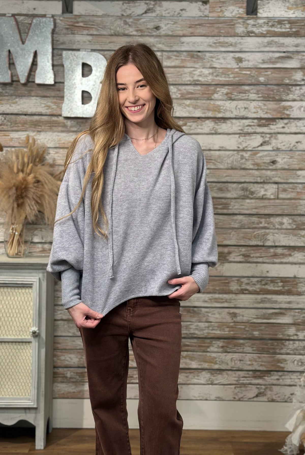 A woman wearing a light gray oversized pullover hoodie with drawstrings and brown pants, standing in front of a rustic wood-paneled wall with decorative letters.