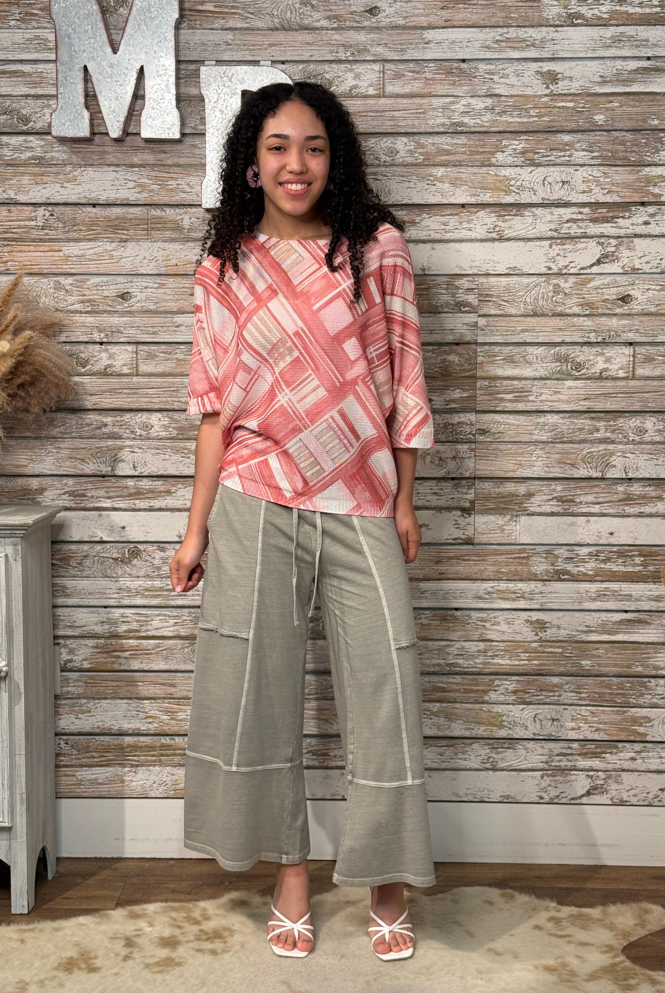 Woman wearing a pink patterned top and beige pants standing against a wooden wall.