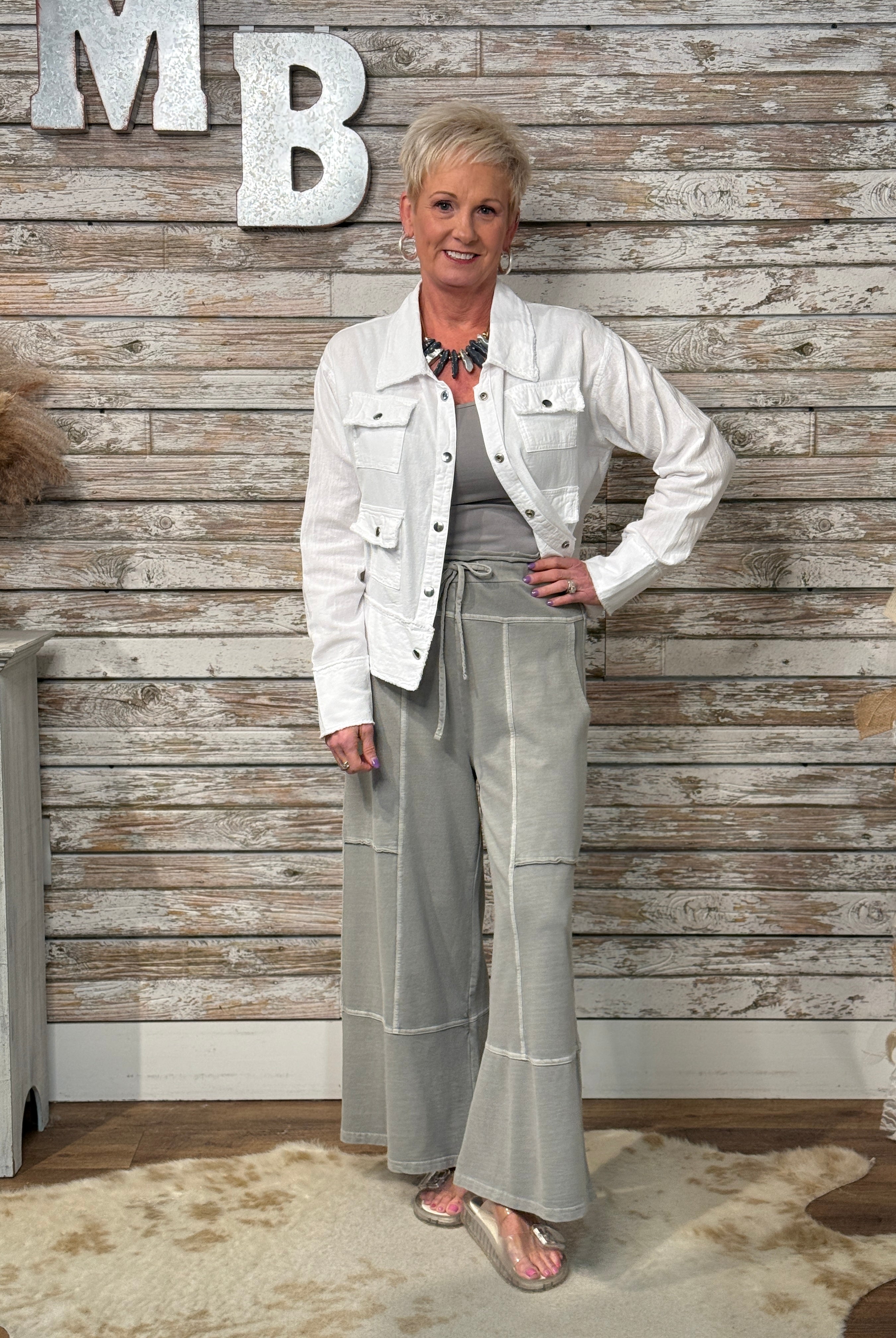 Woman in a white jacket and gray pants standing against a wooden wall with 'MB' letters.
