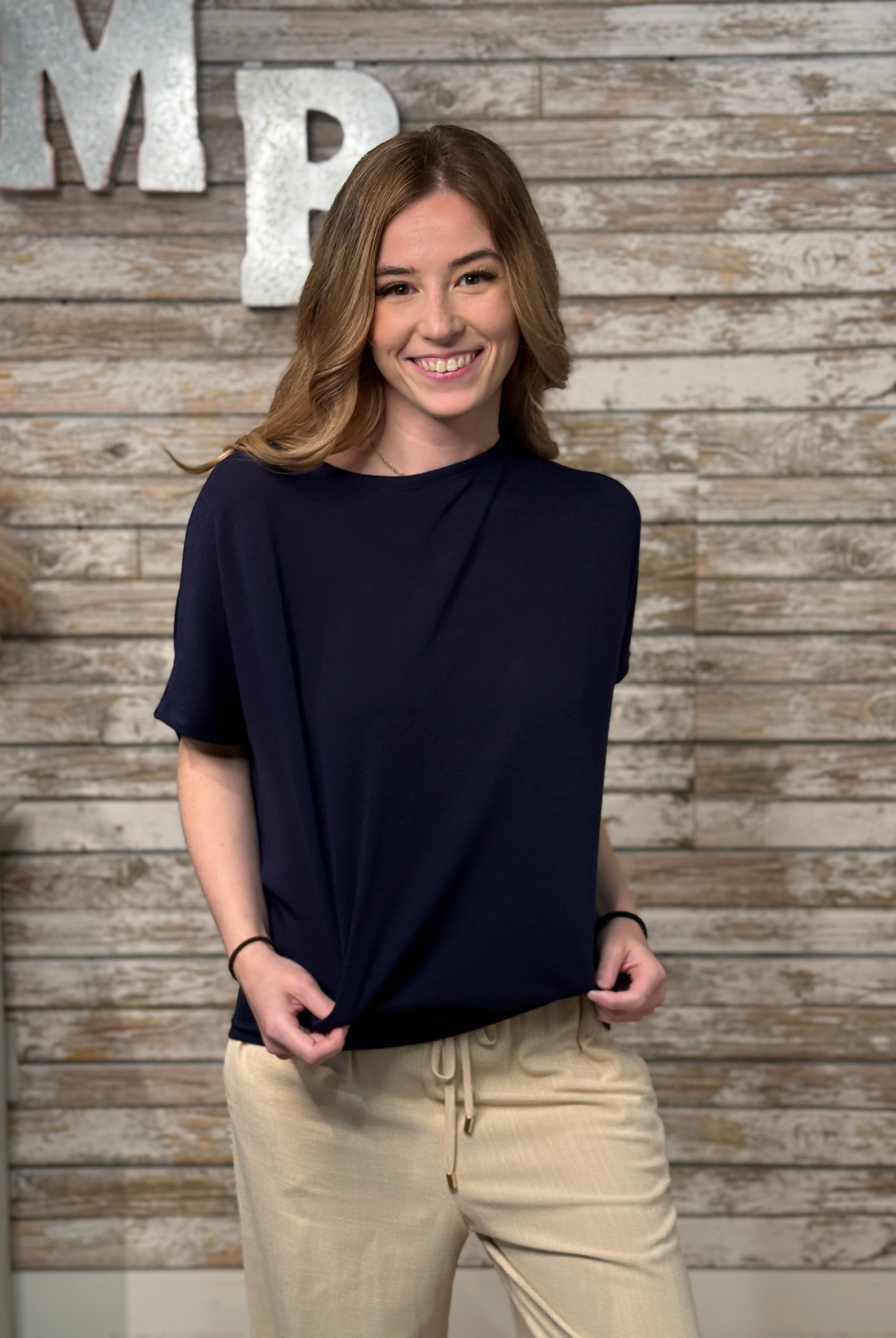 Woman wearing a navy blue top and beige pants standing against a wooden wall with 'MP' letters.