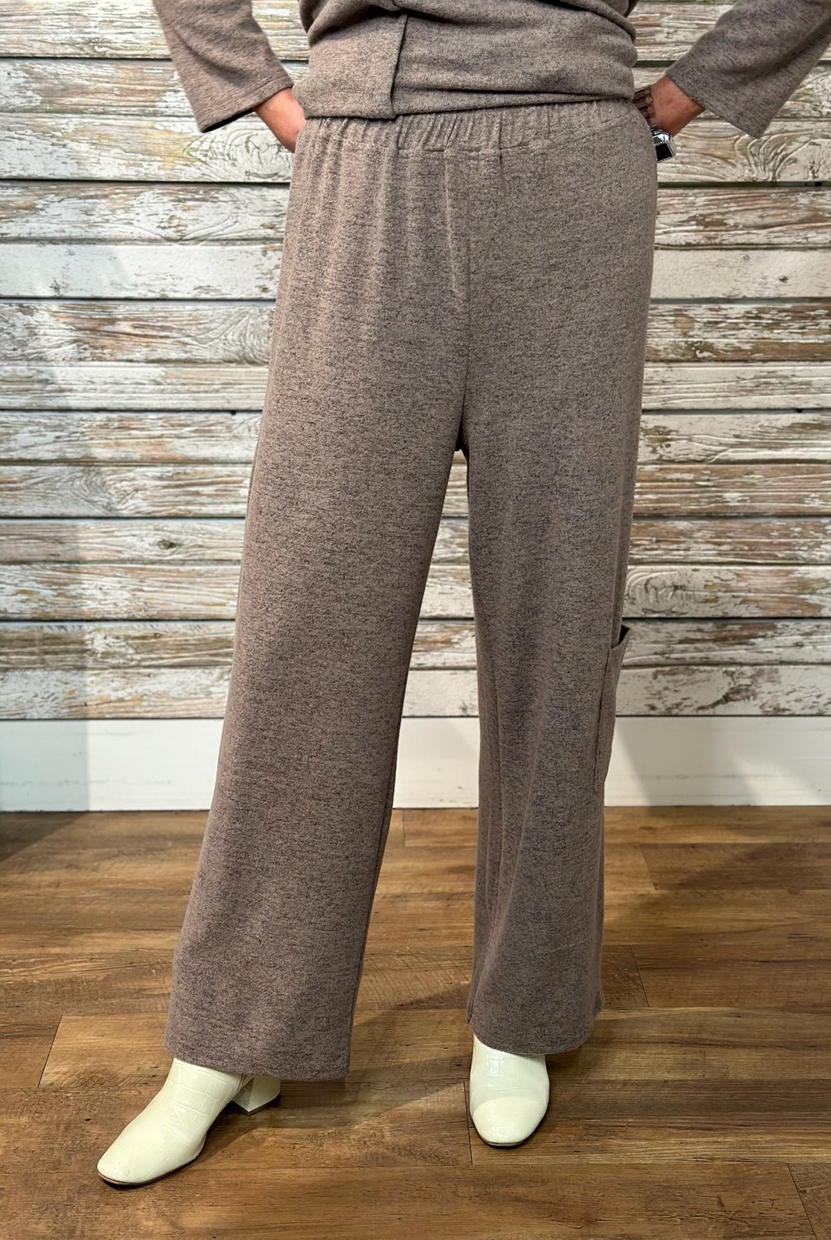 A person wearing loose, wide-leg knit pants in a heather taupe-brown color, paired with a matching top and off-white boots, standing against a rustic whitewashed wood plank wall and wooden floor.