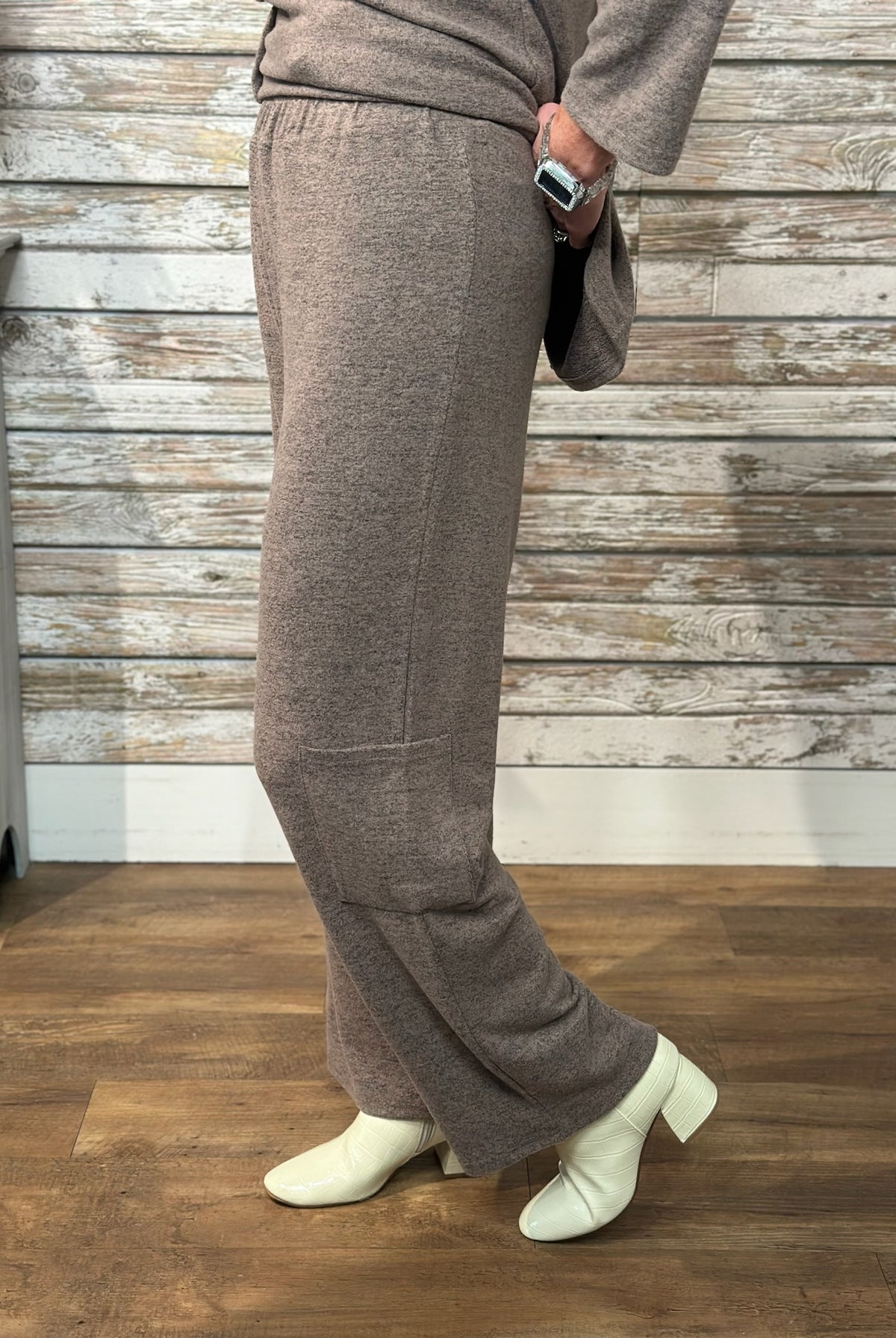 A person wearing a taupe-brown knit long-sleeve top and matching wide-leg pants, paired with off-white ankle boots, standing in front of a rustic whitewashed wood plank wall and wood floor.