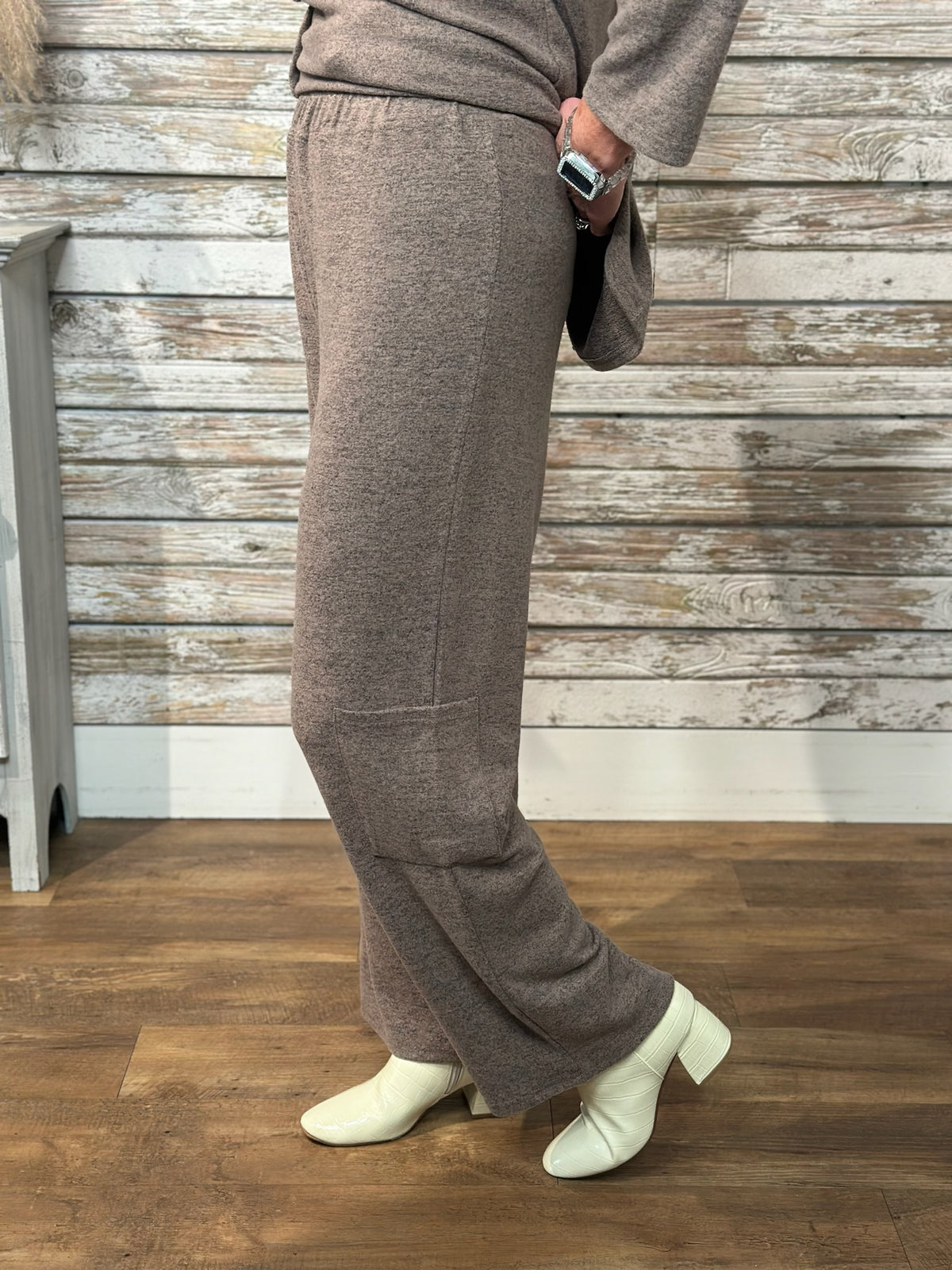A person wearing a taupe-brown knit long-sleeve top and matching wide-leg pants, paired with off-white ankle boots, standing in front of a rustic whitewashed wood plank wall and wood floor.