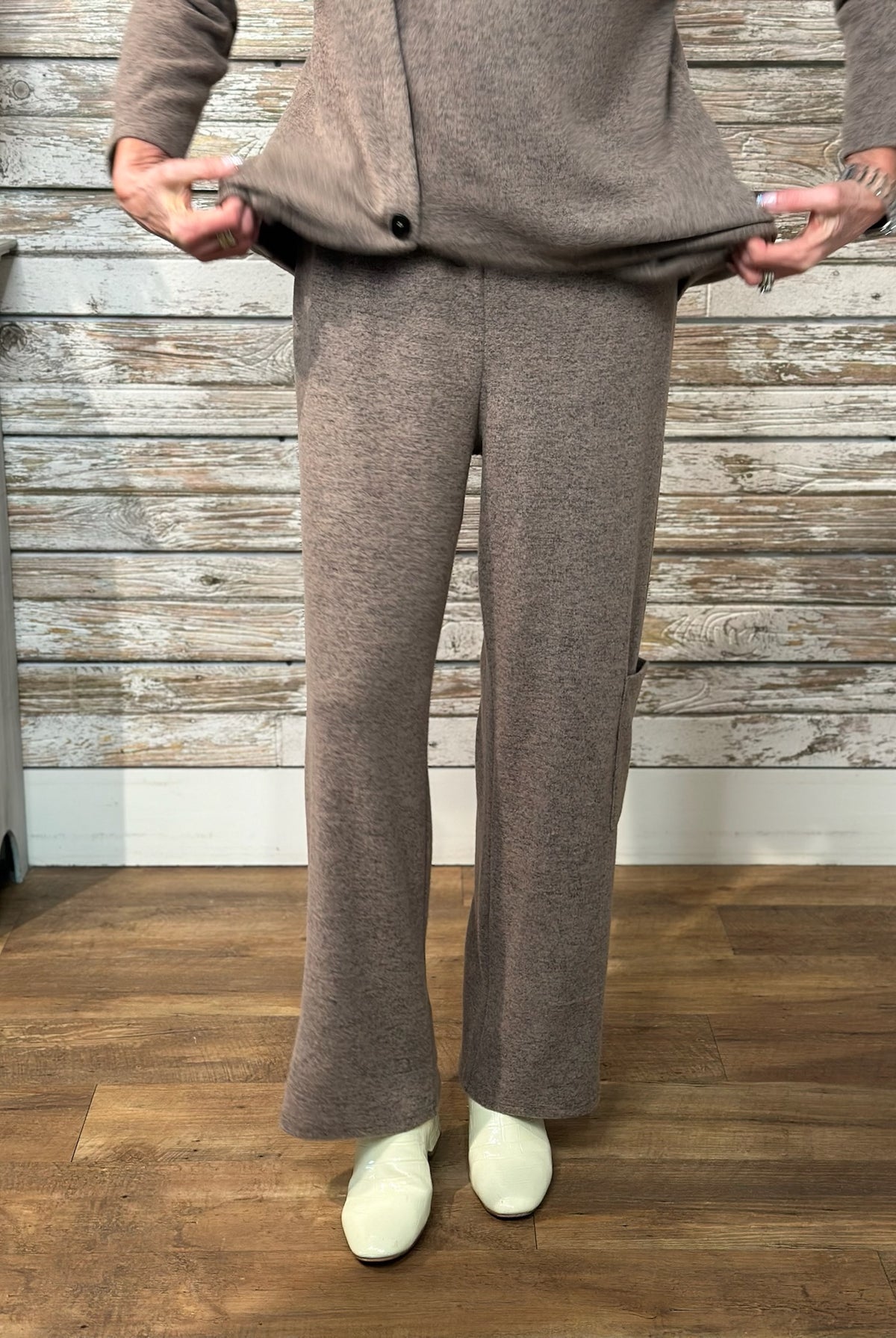 A person modeling a matching taupe knit cardigan and wide-leg pants set, holding the hem of the cardigan to show the fit. The outfit is styled with off-white slip-on shoes, set against a rustic whitewashed wood plank wall and a wood floor.