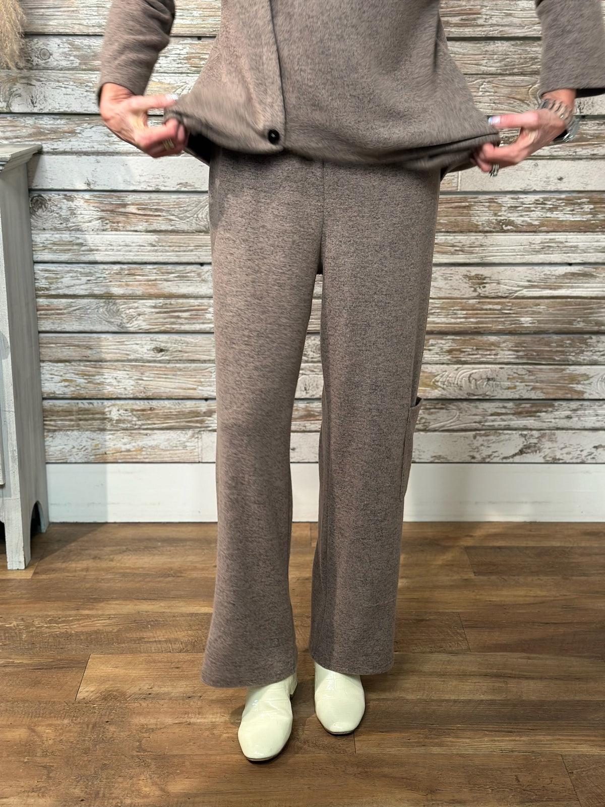 A person modeling a matching taupe knit cardigan and wide-leg pants set, holding the hem of the cardigan to show the fit. The outfit is styled with off-white slip-on shoes, set against a rustic whitewashed wood plank wall and a wood floor.