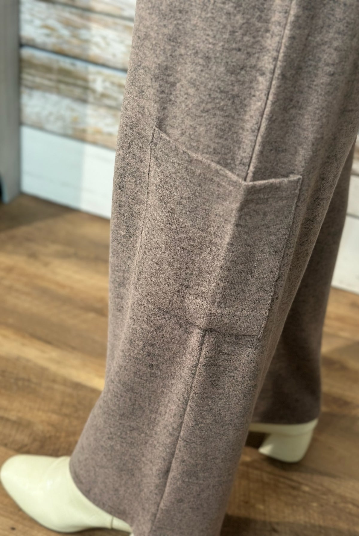 Close-up of a person wearing heather taupe wide-leg trousers with a side pocket, paired with white heeled boots, standing on a wooden floor.